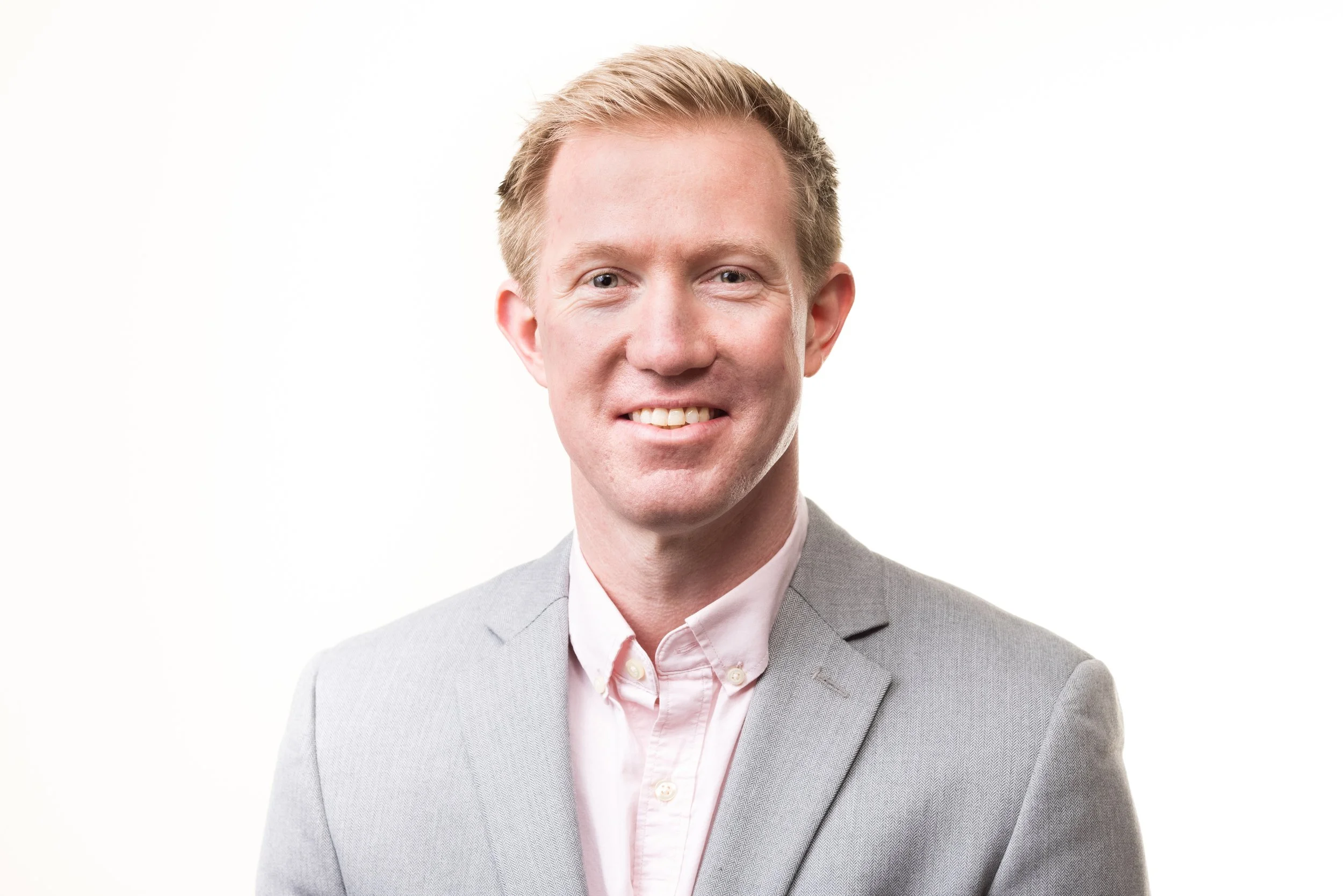 Headshot of a smiling man with light hair wearing a gray suit and white shirt against a white background.