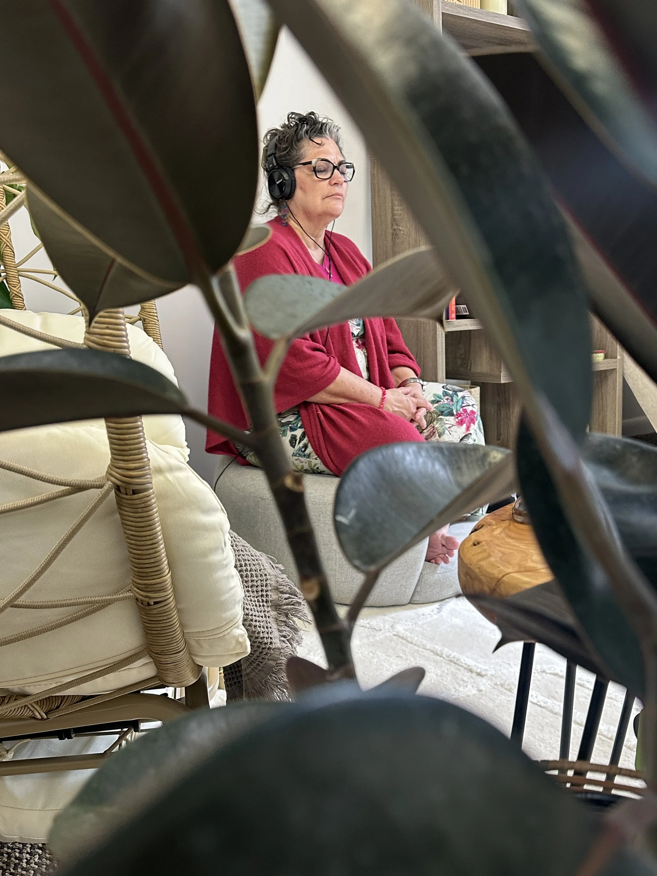 A woman with glasses and curly hair is sitting on a couch, wearing headphones, a red cardigan, and floral pants, with her eyes closed.