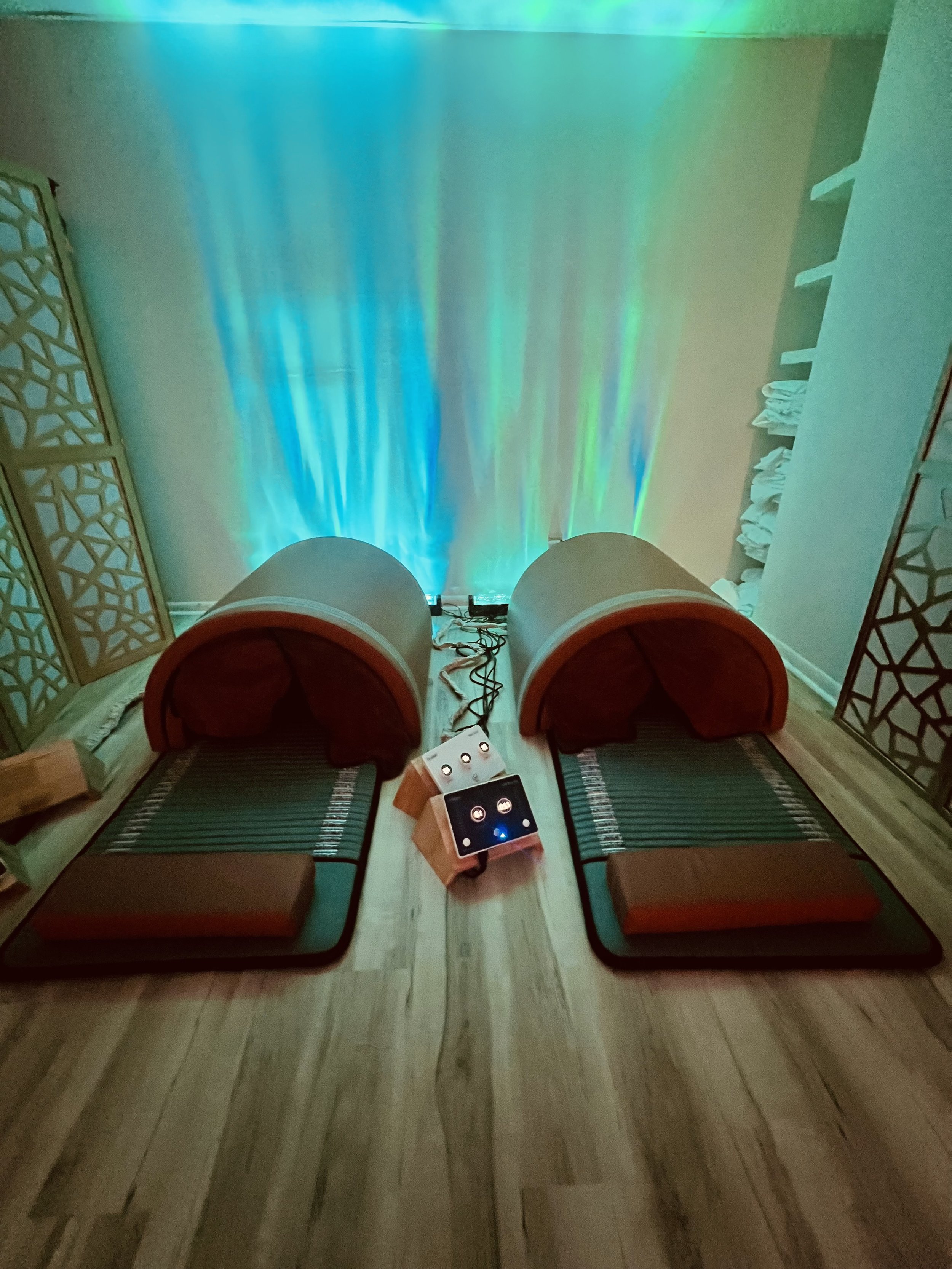 Two identical heated massage pods in a yoga or massage room, with colorful LED lighting on the wall behind them, surrounded by decorative room dividers and white towels stacked on shelves.