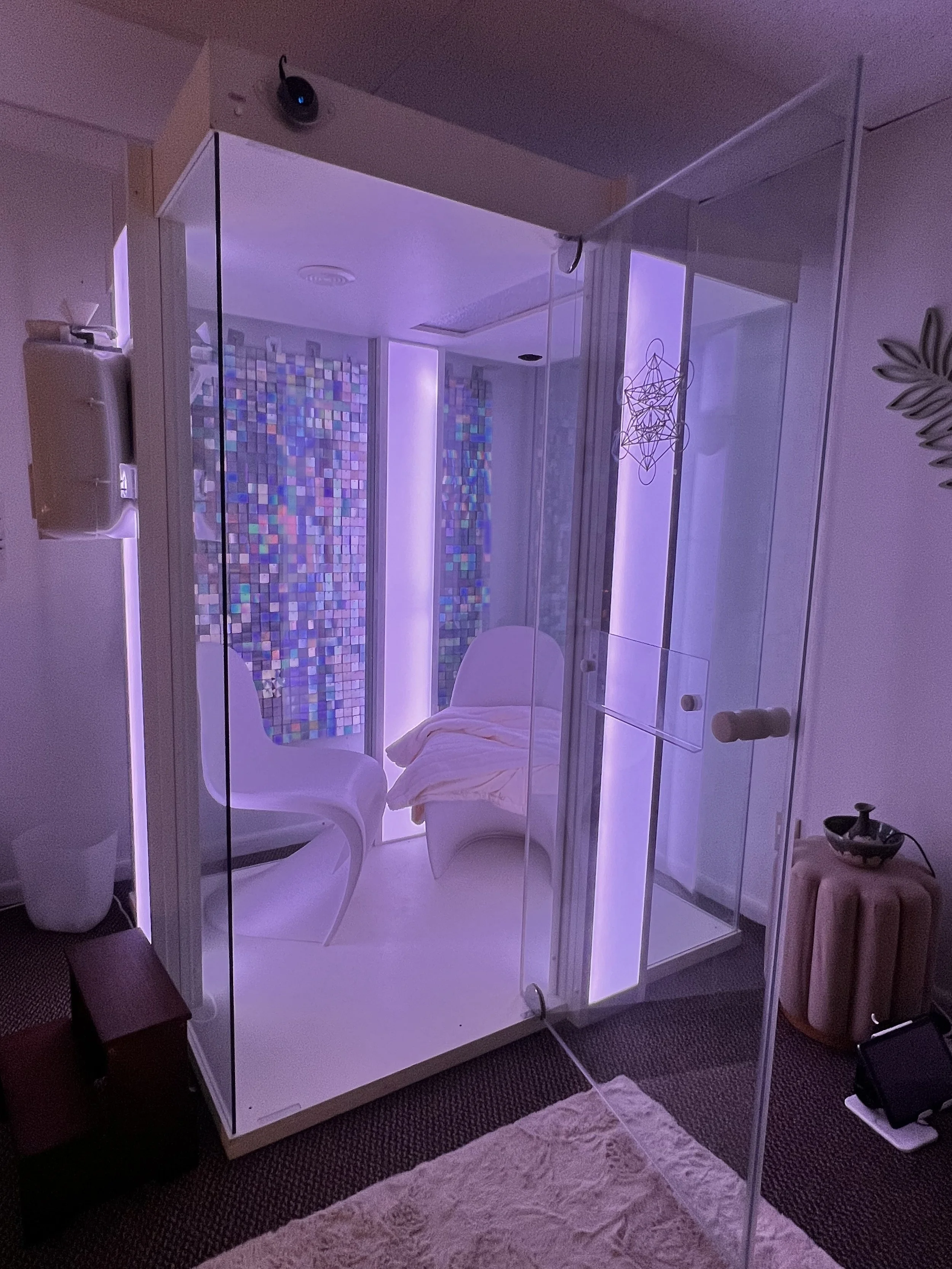A small transparent glass booth with purple lighting, two white modern chairs, a blanket draped over one chair, and a small device mounted on the outside wall.