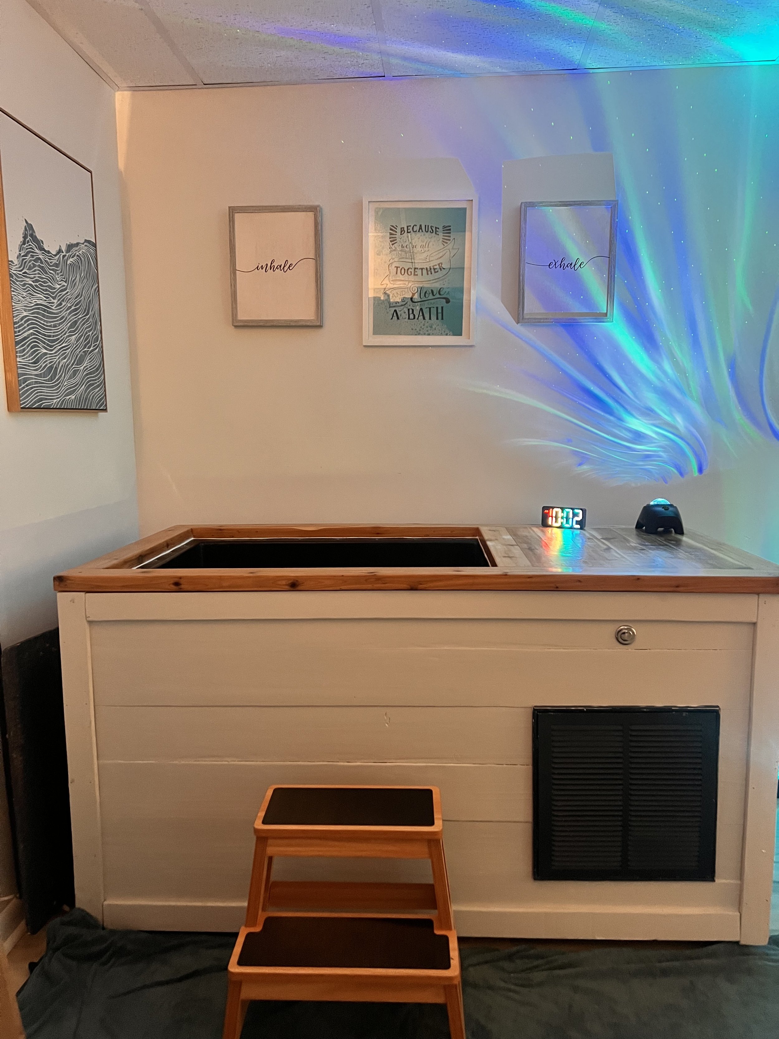 Indoor spa room with cold plunge, wooden steps, towels on a ladder shelf, a small side table, a decorative room divider, and blue ambient lighting.