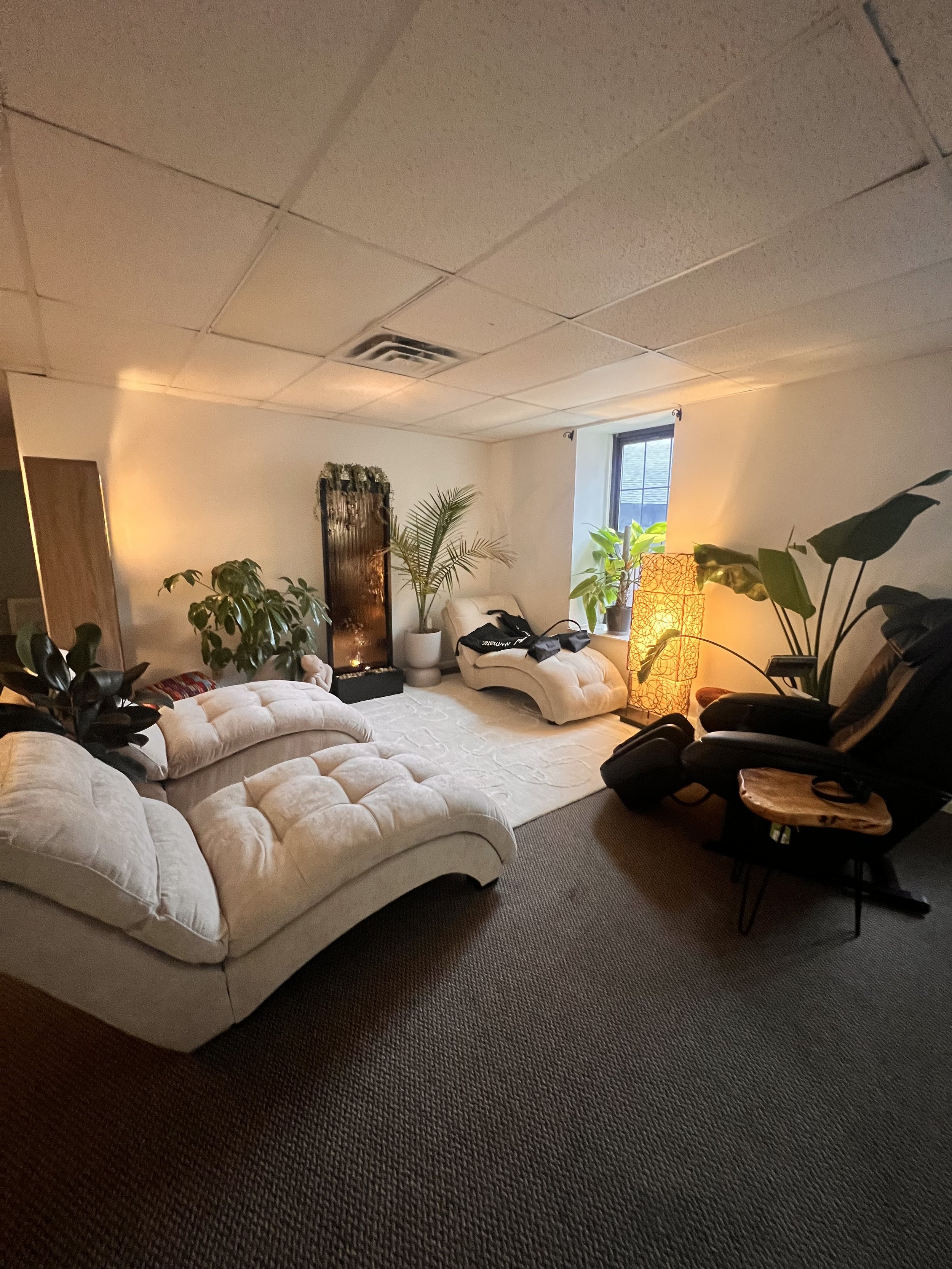 A cozy room with a beige sectional sofa, a black massage chair, a wooden side table, various potted plants near a window, a standing lamp with warm yellow light, and a wall-mounted water feature with plants and lights.