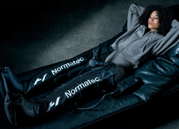 Woman resting on a black massage chair with Normatec compression leg sleeves and remote control.