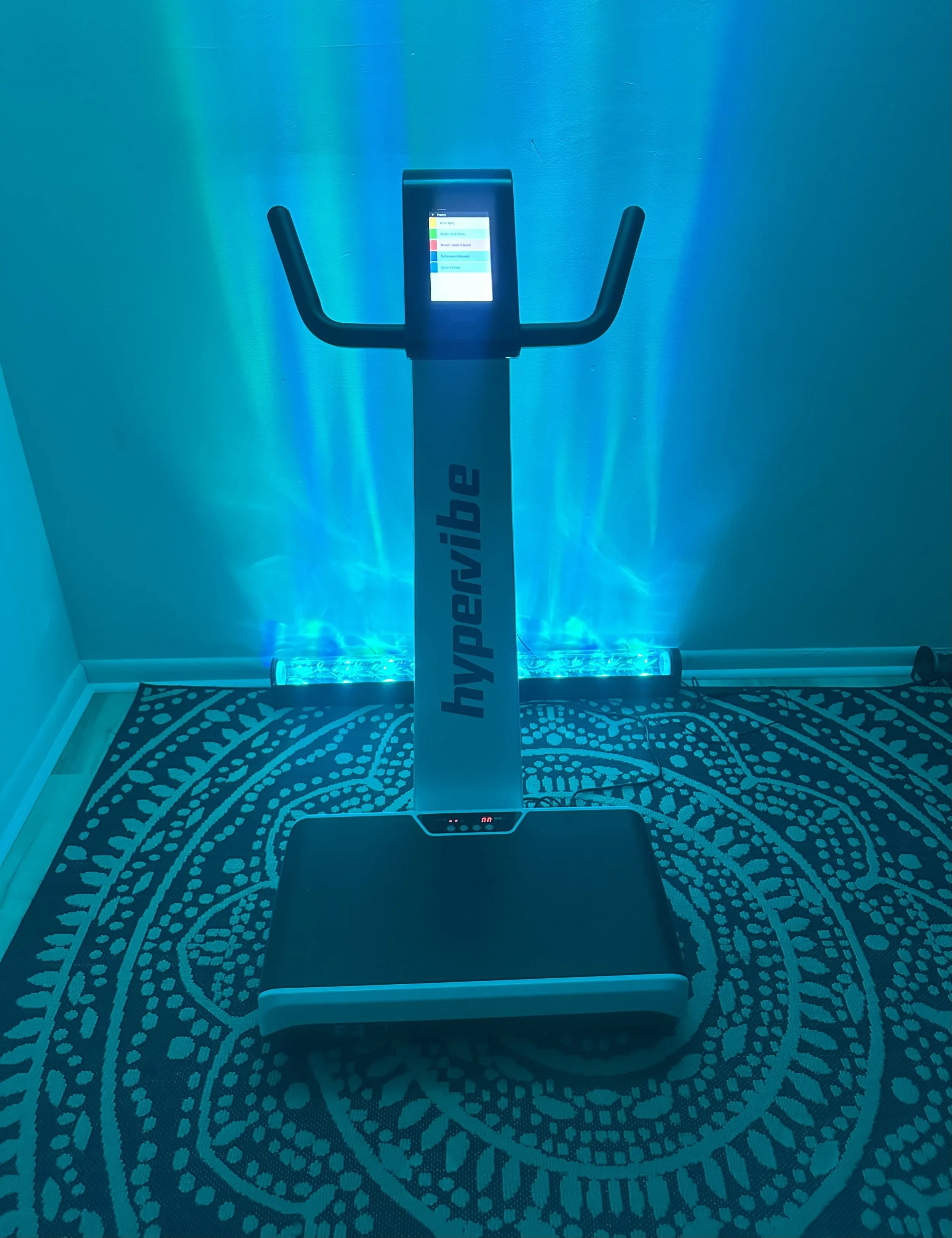 Hypervibe vibration platform machine in a room with blue lighting, on a patterned rug.
