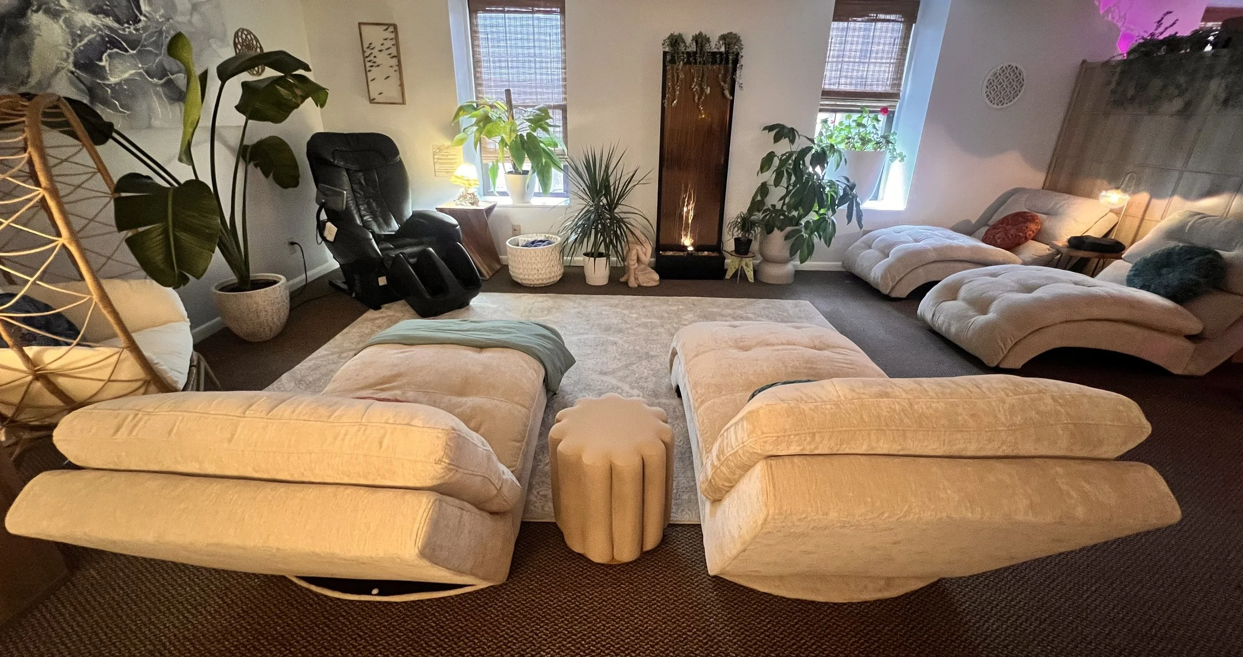 A cozy room with a beige sectional sofa, a black massage chair, a wooden side table, various potted plants near a window, a standing lamp with warm yellow light, and a wall-mounted water feature with plants and lights.