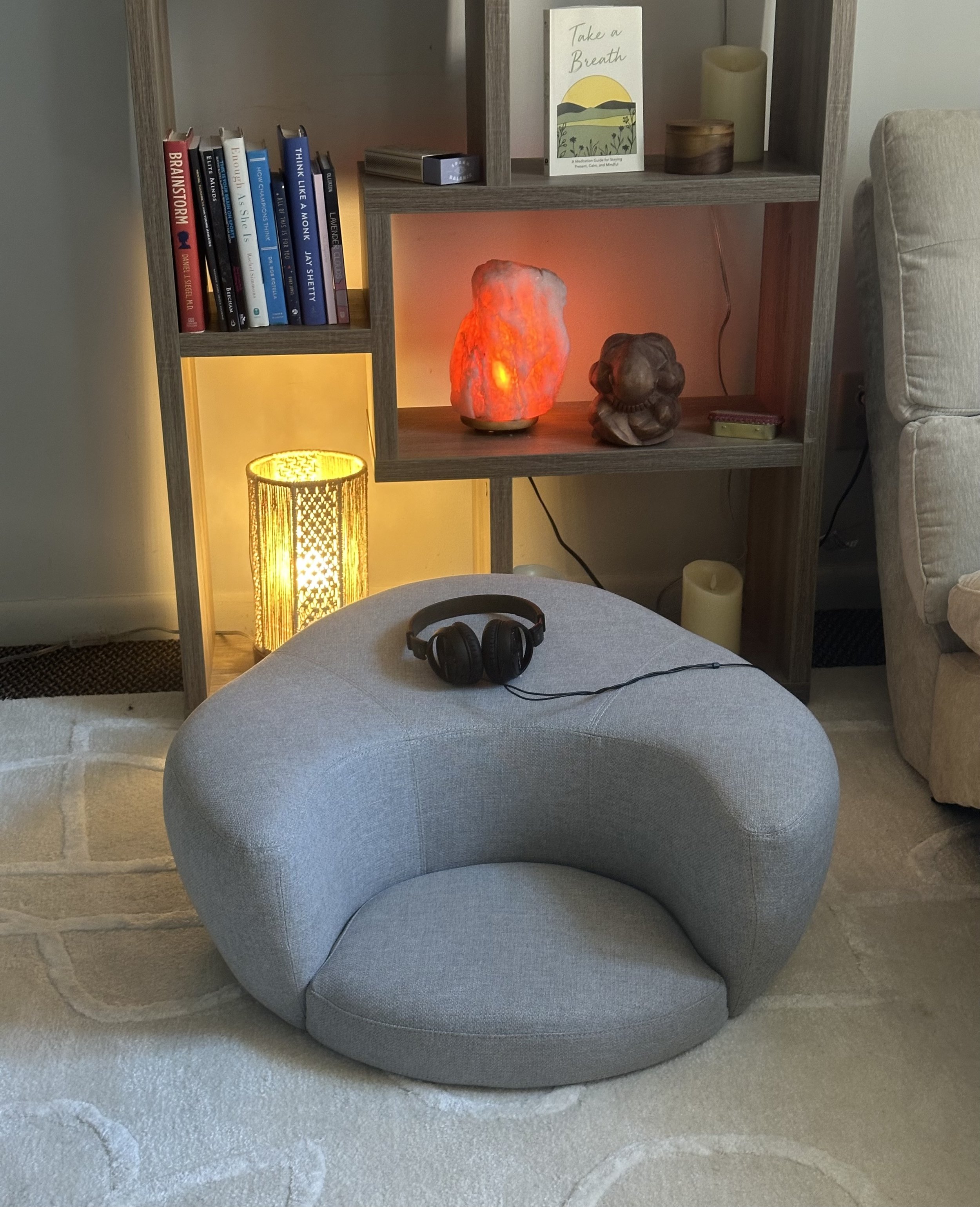A cozy room corner with a gray round armchair, a pair of black headphones on it, a wooden bookshelf with books, salt lamps, and a decorative rock lamp emitting warm light, a yellow wicker lamp, a beige upholstered sofa, and a small rug.