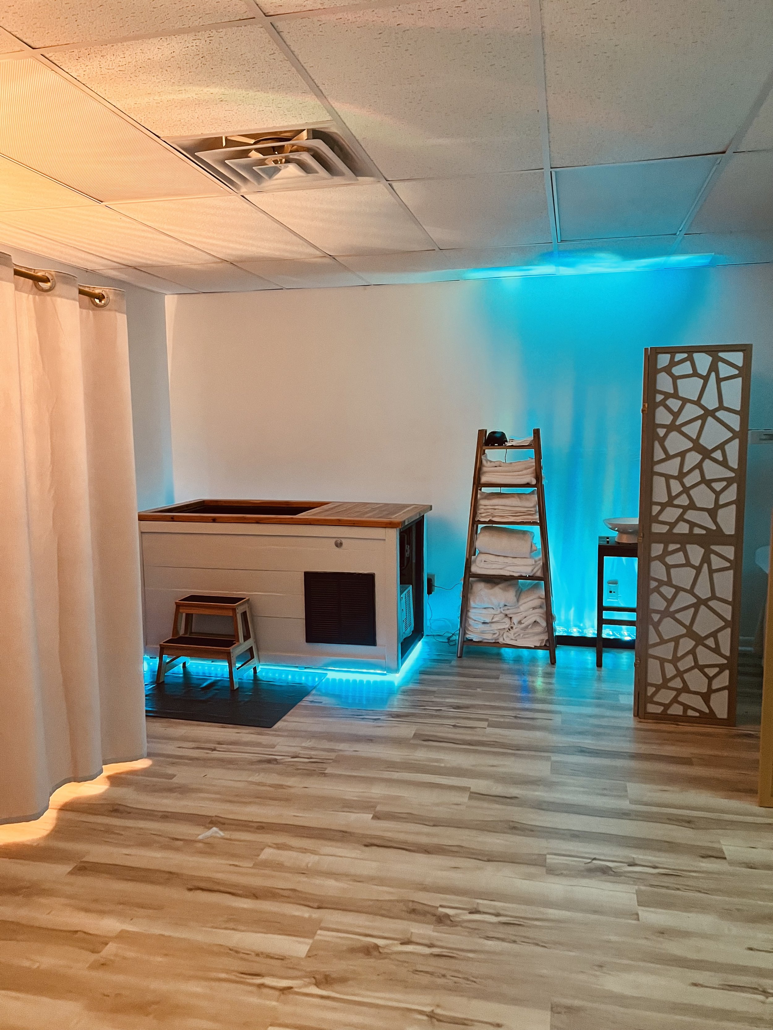 Indoor spa room with cold plunge, wooden steps, towels on a ladder shelf, a small side table, a decorative room divider, and blue ambient lighting.