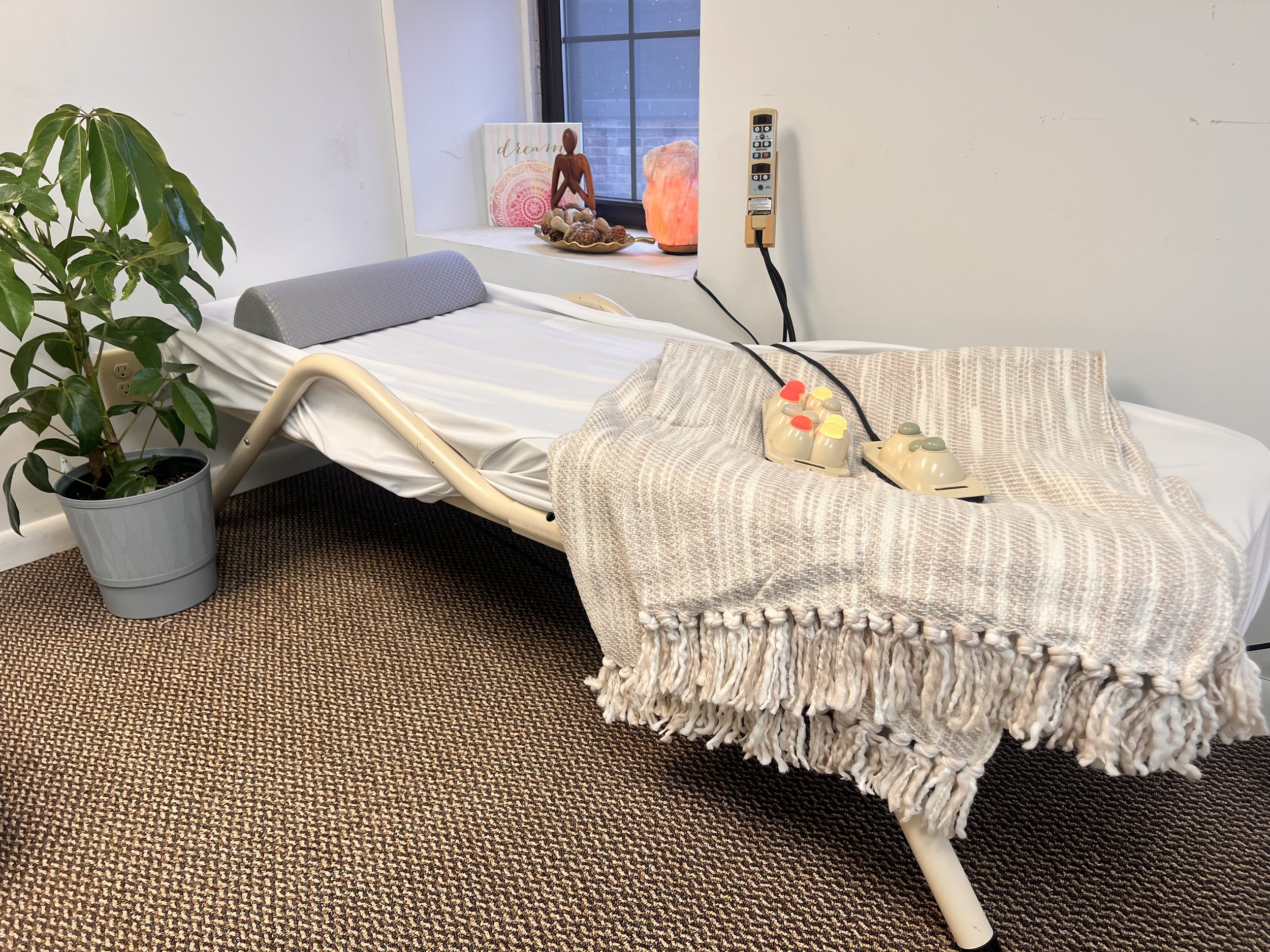 Hospital bed with a gray pillow, a potted plant, a window with decorative items on the sill, including a salt lamp, a figurine, and a book, and a beige blanket with medical devices on top.