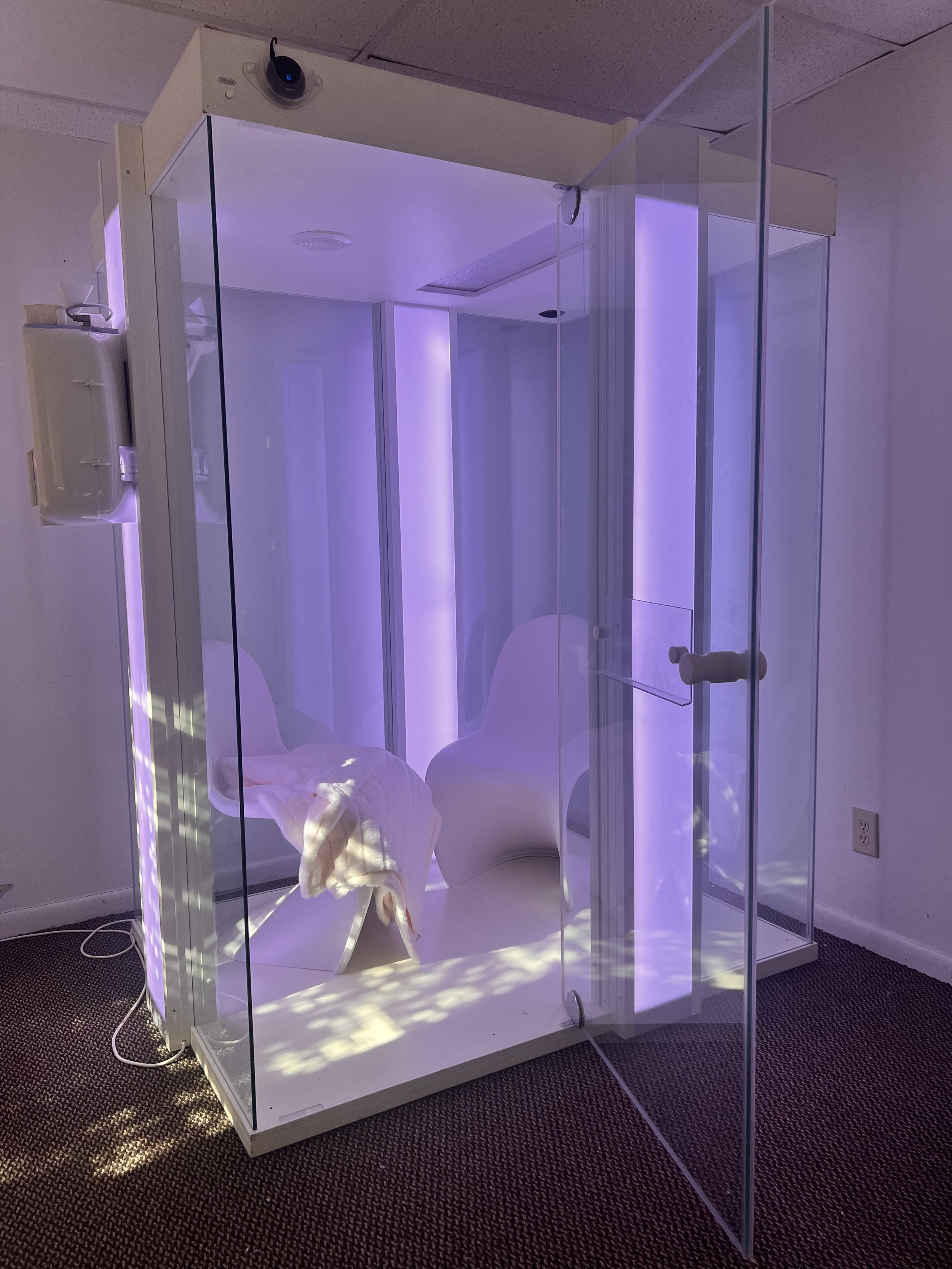 A small transparent glass booth with purple lighting, two white modern chairs, a blanket draped over one chair, and a small device mounted on the outside wall.