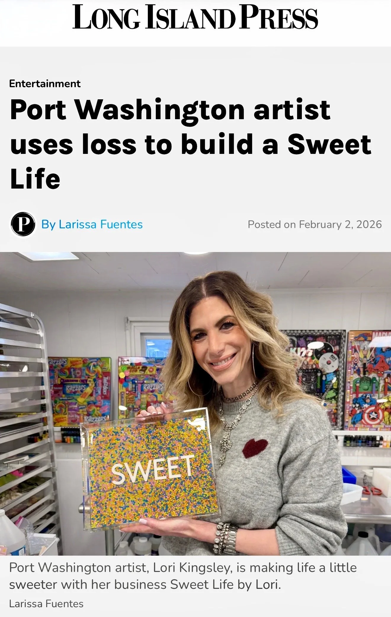 Lori Kingsley, a Port Washington artist featured in the Long Island Press, holding a colorful, decorated serving tray with the word 'SWEET' on it. She is smiling and wearing jewelry, in a room with shelves of art.
