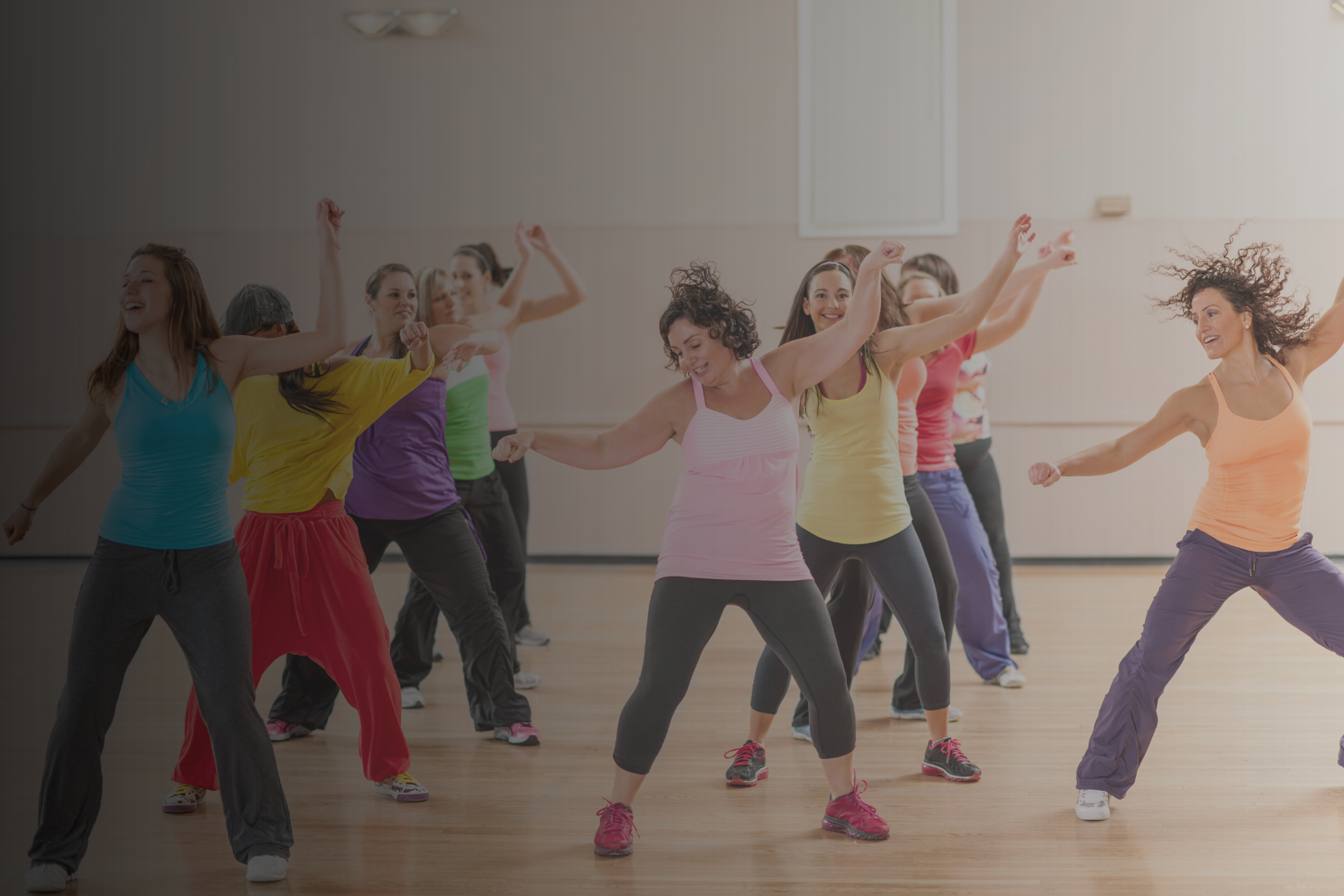A group of women dancing and exercising in a dance studio or gym, wearing colorful athletic clothing.