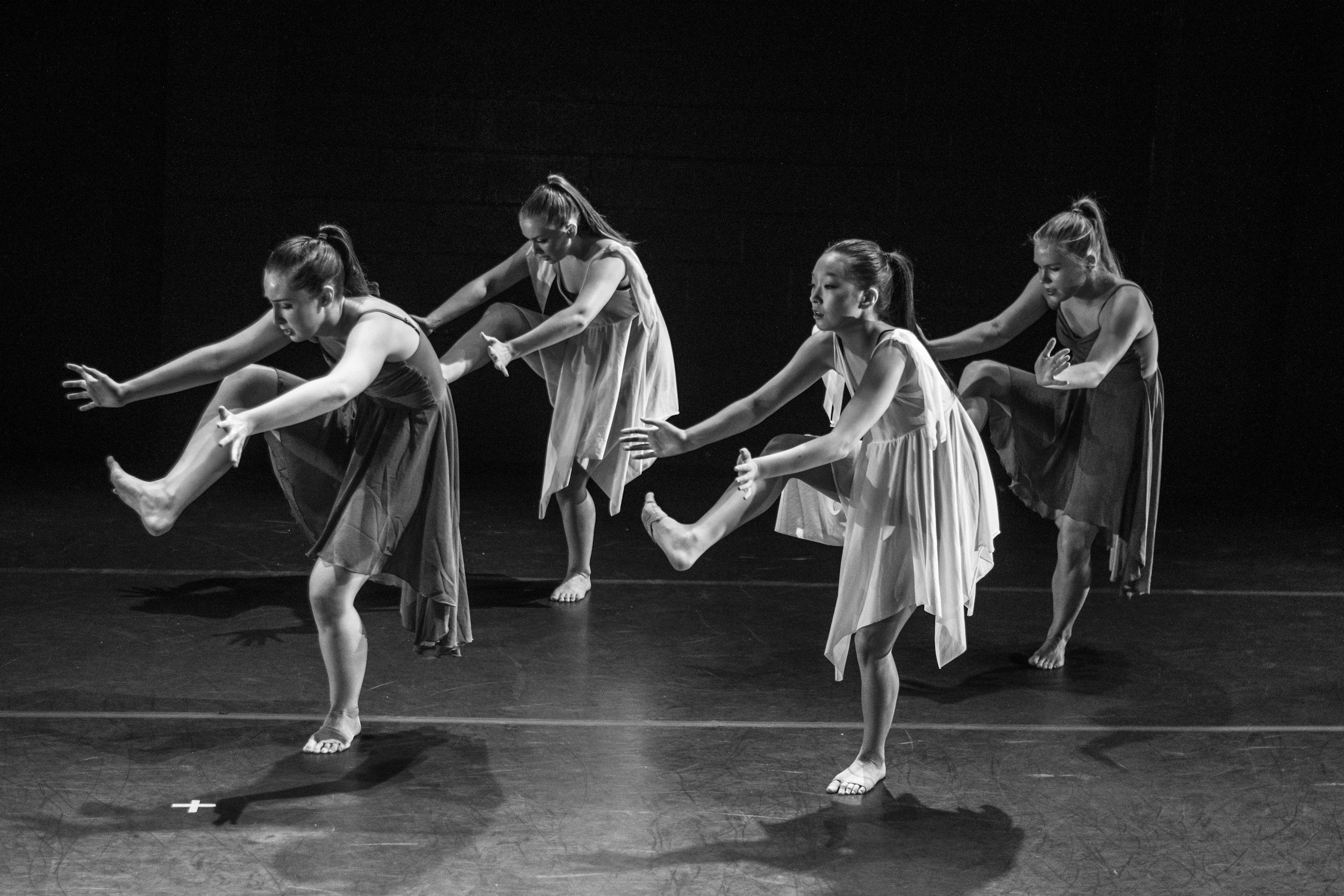 Four young female dancers in sleeveless dresses performing a synchronized dance move on a dark stage, all raising one leg and extending their arms forward.