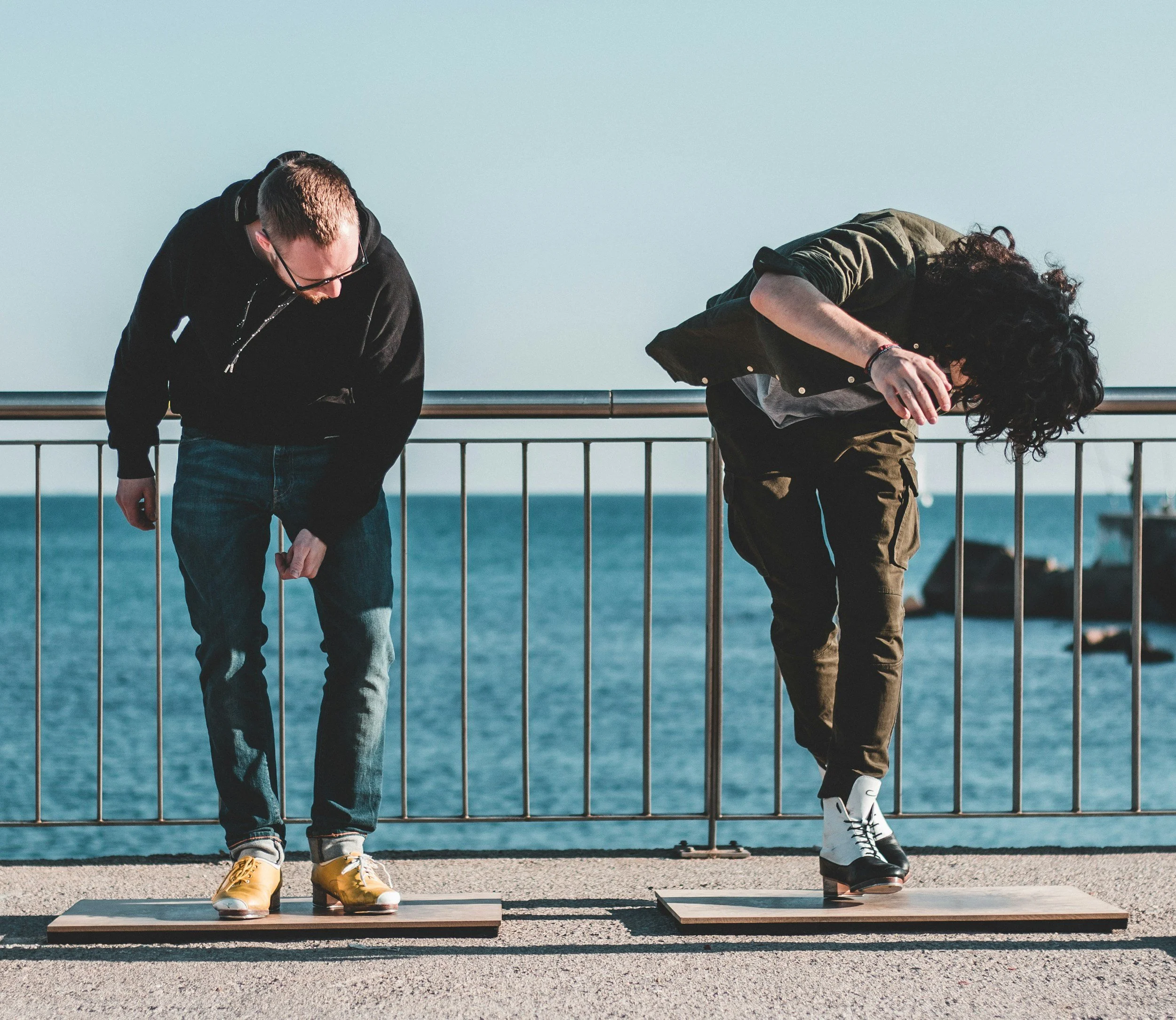 Two men are standing on wooden platforms near a body of water, both leaning forward with their heads down, wearing ice skates, and dressed in casual clothing.
