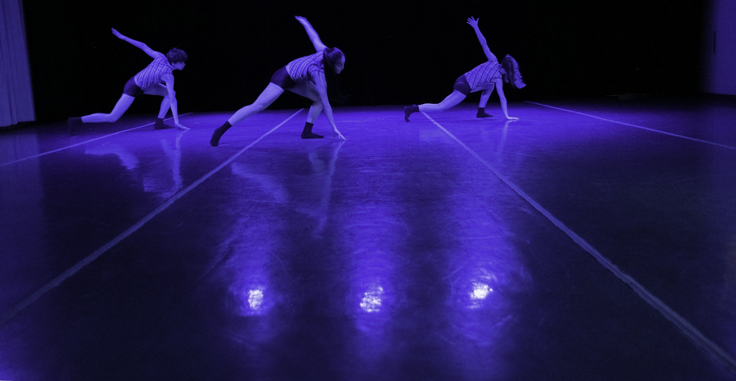 Three dancers perform on a dark stage illuminated by blue lighting, wearing matching striped shirts and shorts, in various dynamic poses.