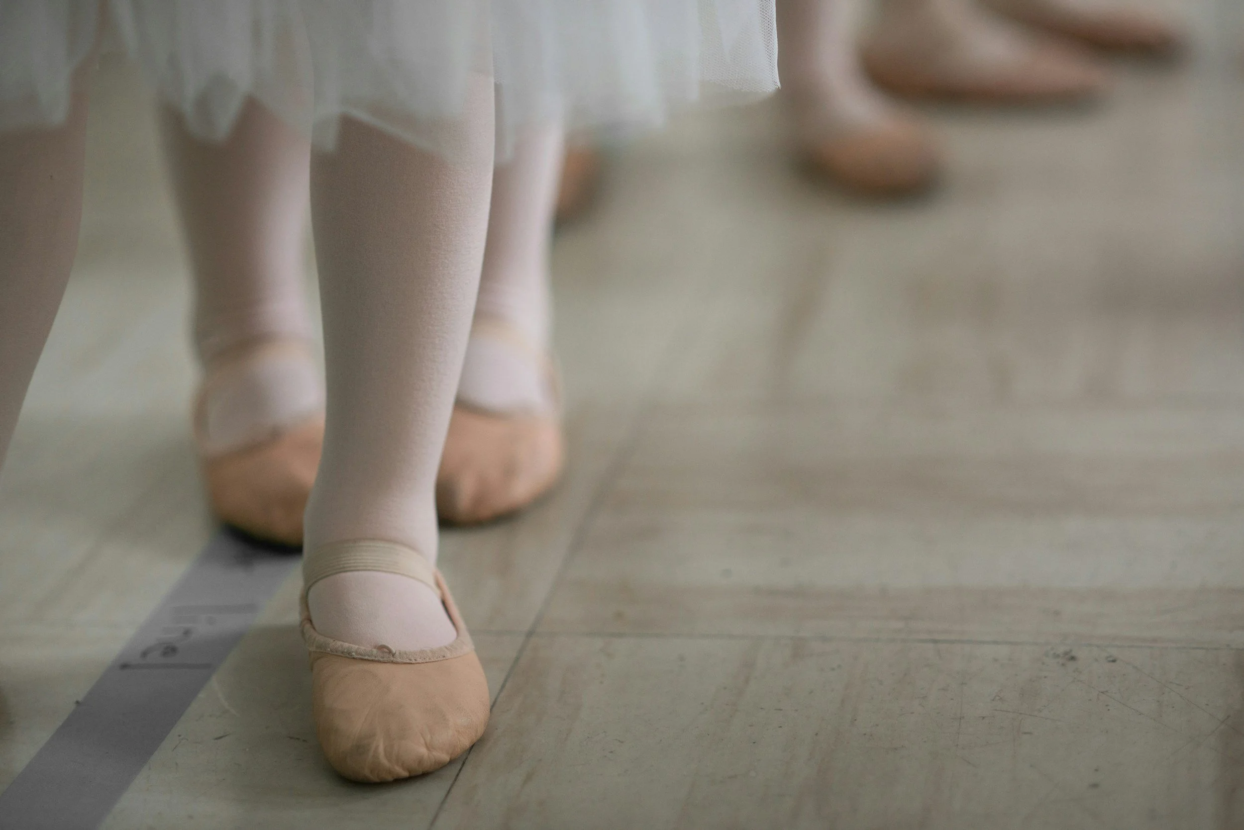 Close-up of young ballerinas' feet in ballet slippers and tights, standing on a dance floor.