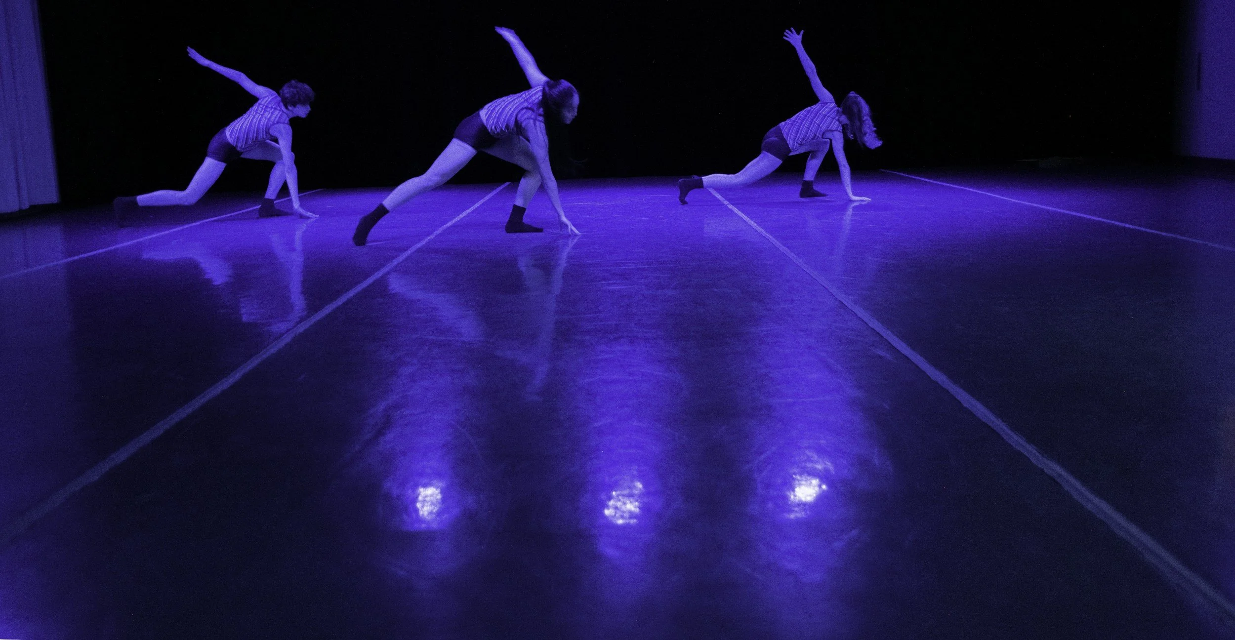 Three dancers in striped shirts and shorts performing on a dark stage with blue lighting, posing in a stretch on the floor.