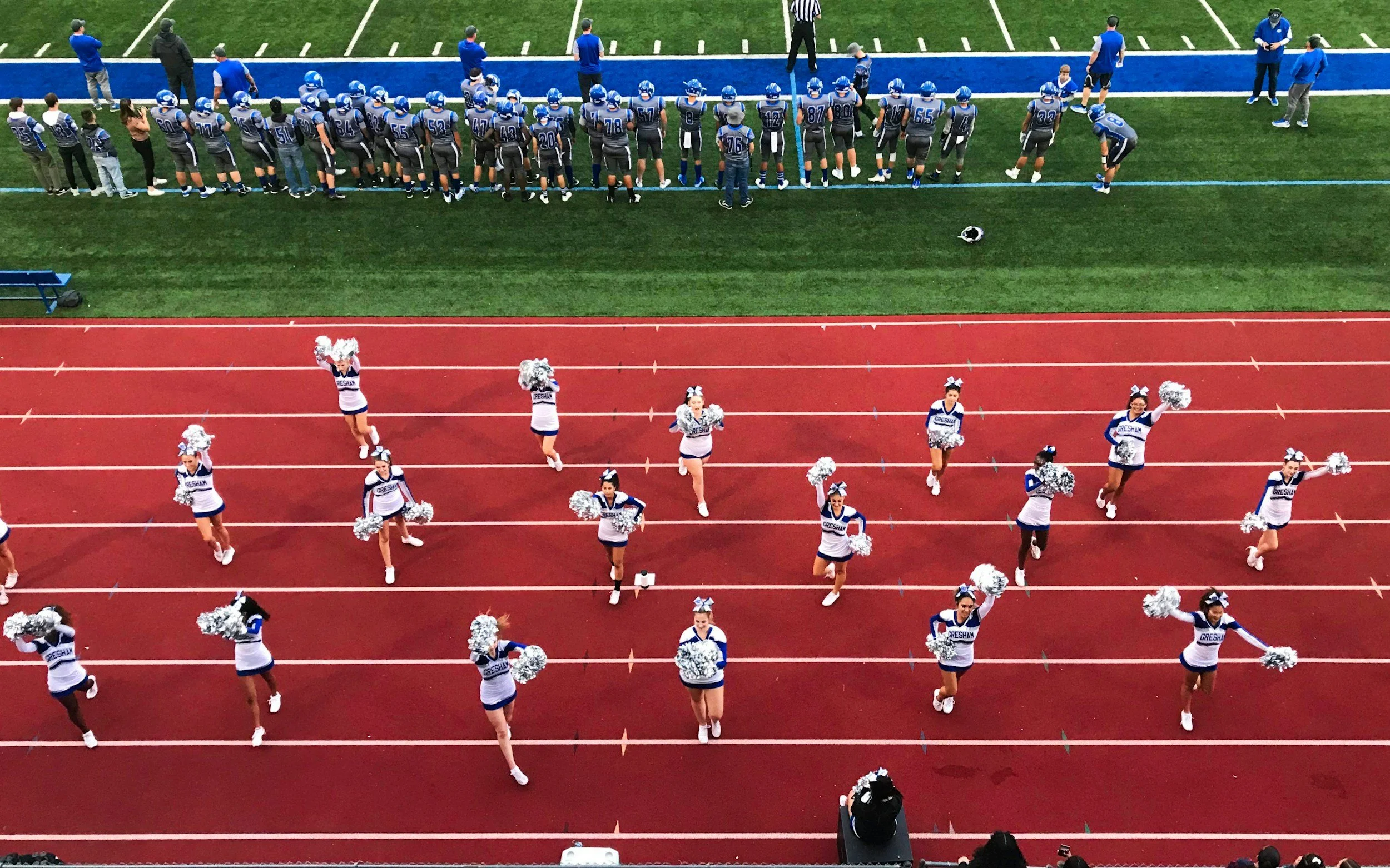 Cheerleaders performing on a red track with football players in the background.