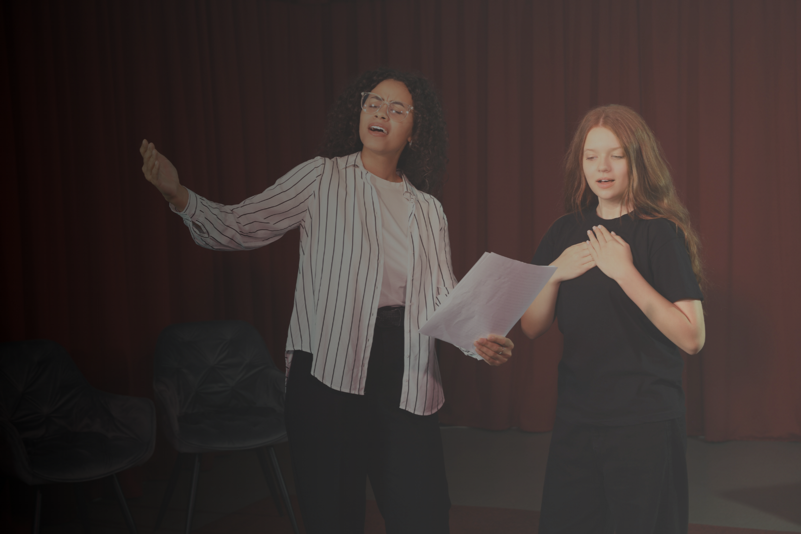 A woman singing passionately with a sheet of paper in her hand, and a young girl with her hand on her chest singing or reciting, in front of a red curtain.