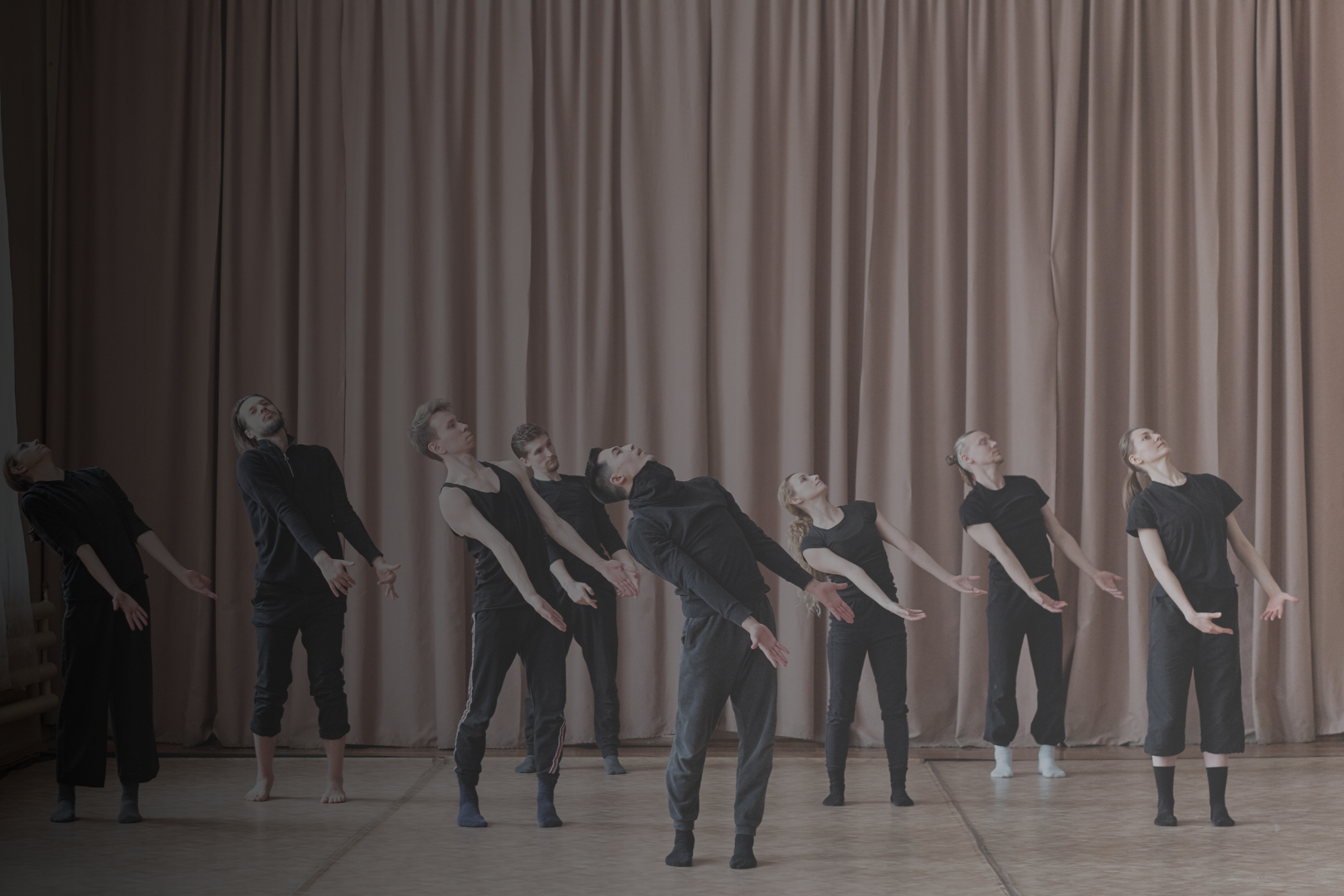 Group of nine dancers in black clothing practicing on stage in front of beige curtain, performing a synchronized dance move with arms extended and heads tilted back.