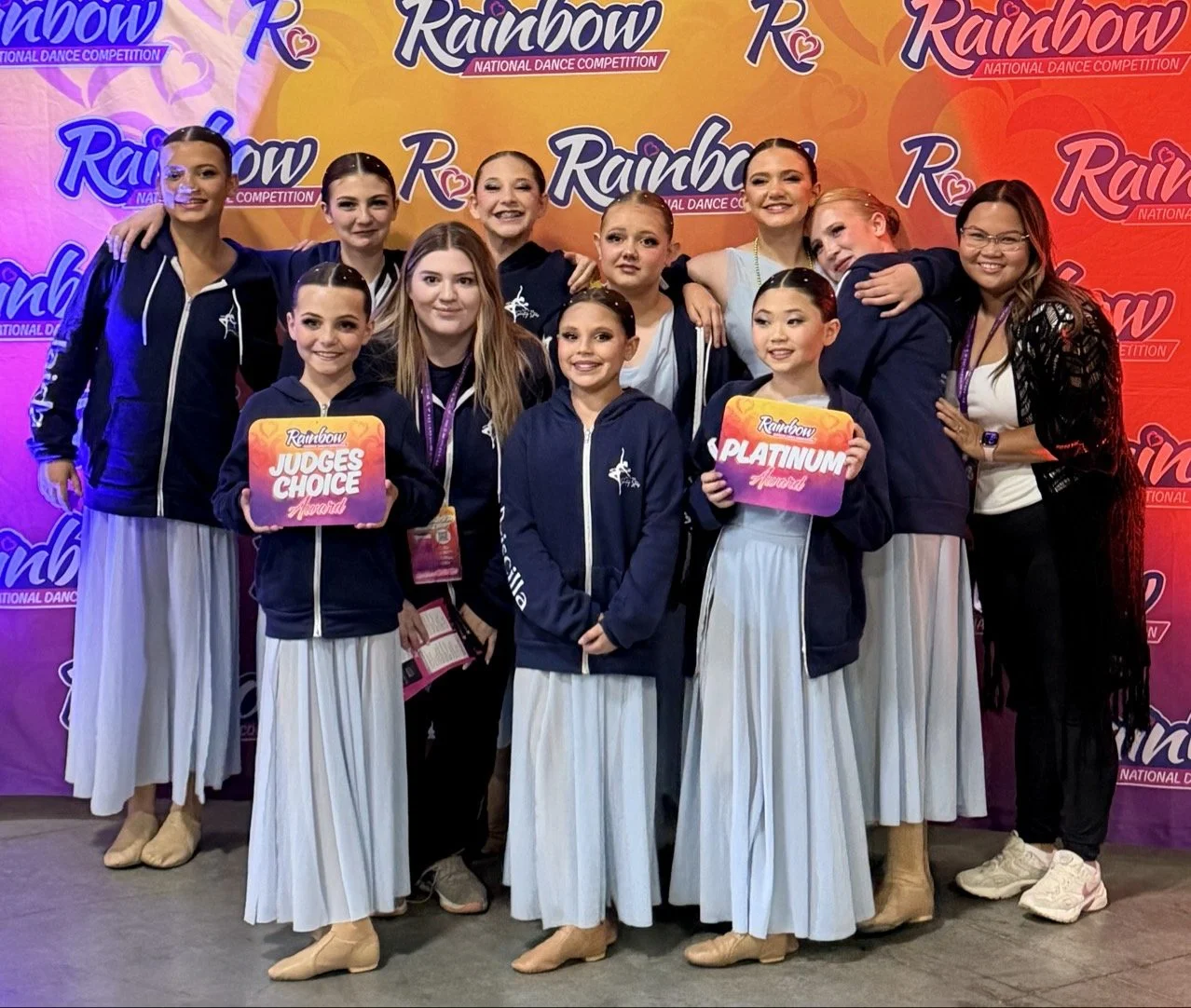 Group of young female dancers in white skirts and dark hoodies, standing together at a dance competition, holding awards and medals, with colorful backdrop featuring 'Rainbow' and 'National Dance Competition' logos.