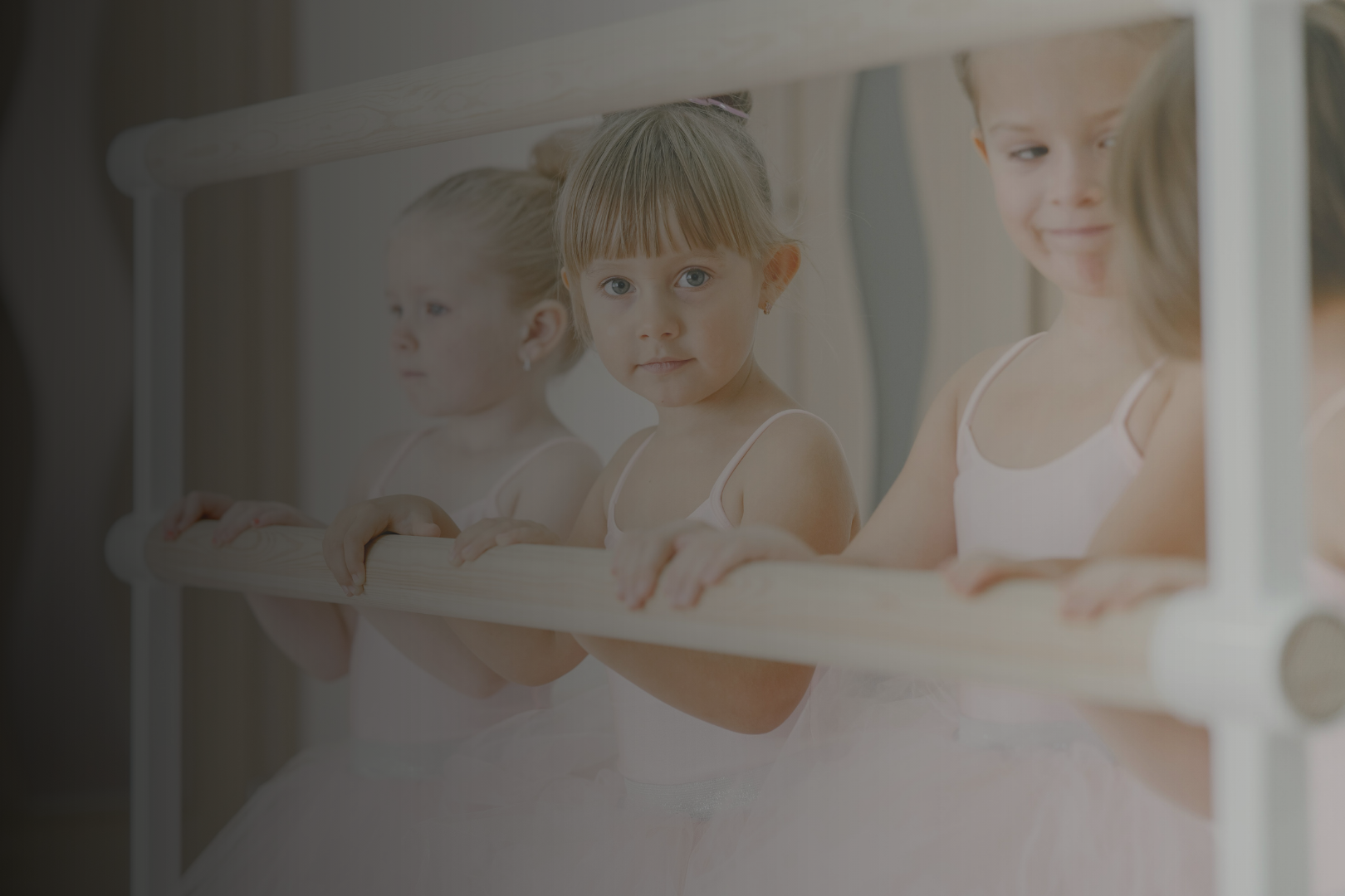 Three young girls dressed in ballet tutus holding onto a ballet barre, looking at the camera.