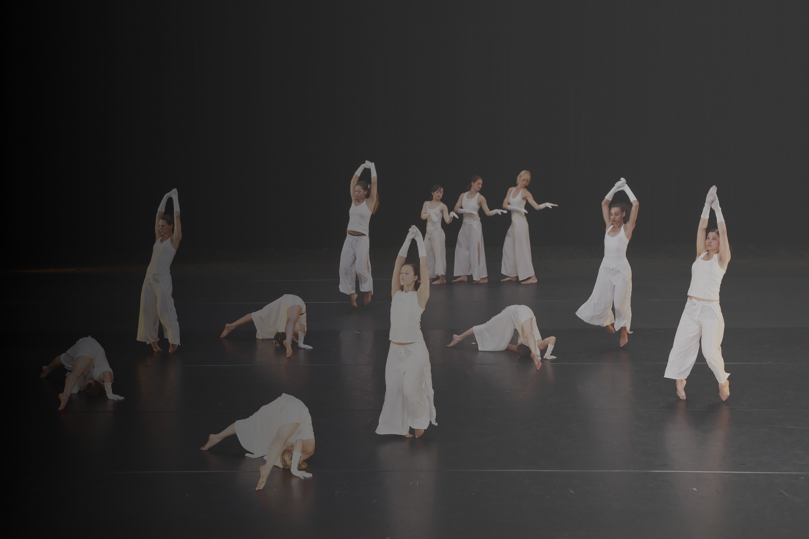Group of dancers performing a choreographed dance on stage, wearing white costumes with gloves, in various poses, with some on their hands and knees and others standing and stretching.