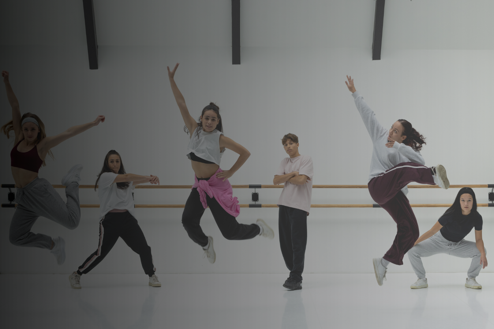 Group of six dancers practicing in a dance studio with ballet barres on the wall. They are in various dance poses, some jumping and some crouching, dressed in casual workout clothes.