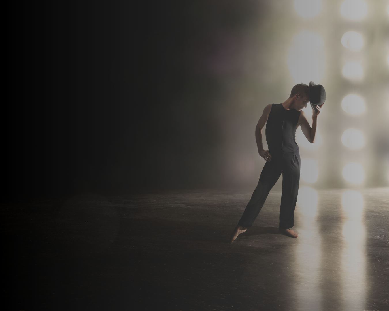A dancer on stage with a dramatic lighting effect, holding a black hat in one hand and dressed in black, barefoot.