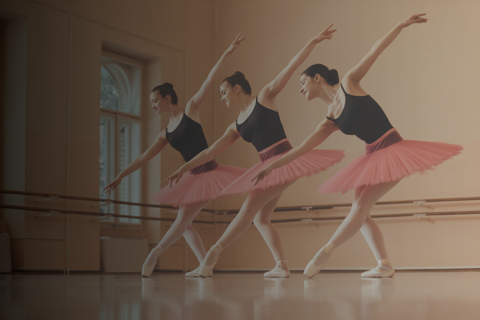 Three ballerinas in black leotards and pink tutus practicing ballet in a dance studio with large windows and wooden ballet barre.