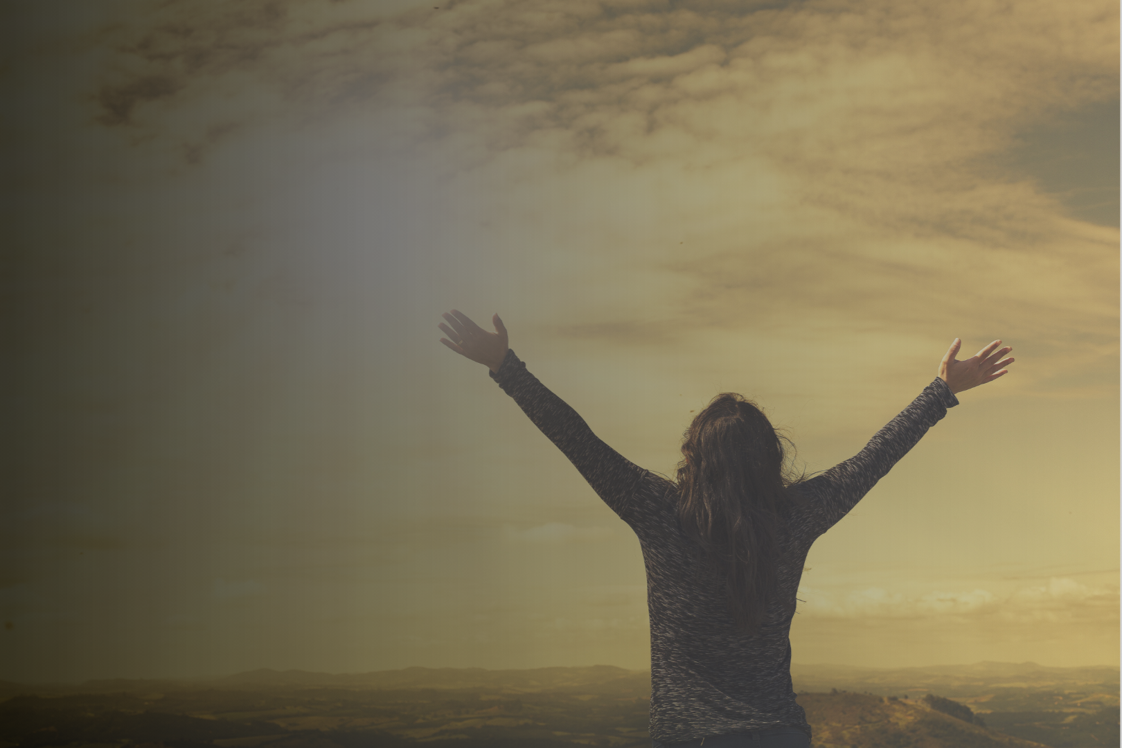 A person standing outdoors with arms raised towards the sky during sunset, with cloudy sky and distant mountains.