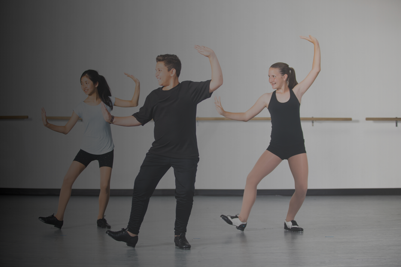 Three dancers in a dance studio practicing ballet or contemporary dance, with two women and one man. All are in casual dancewear, with the man in the center and the women on either side, performing dance poses.