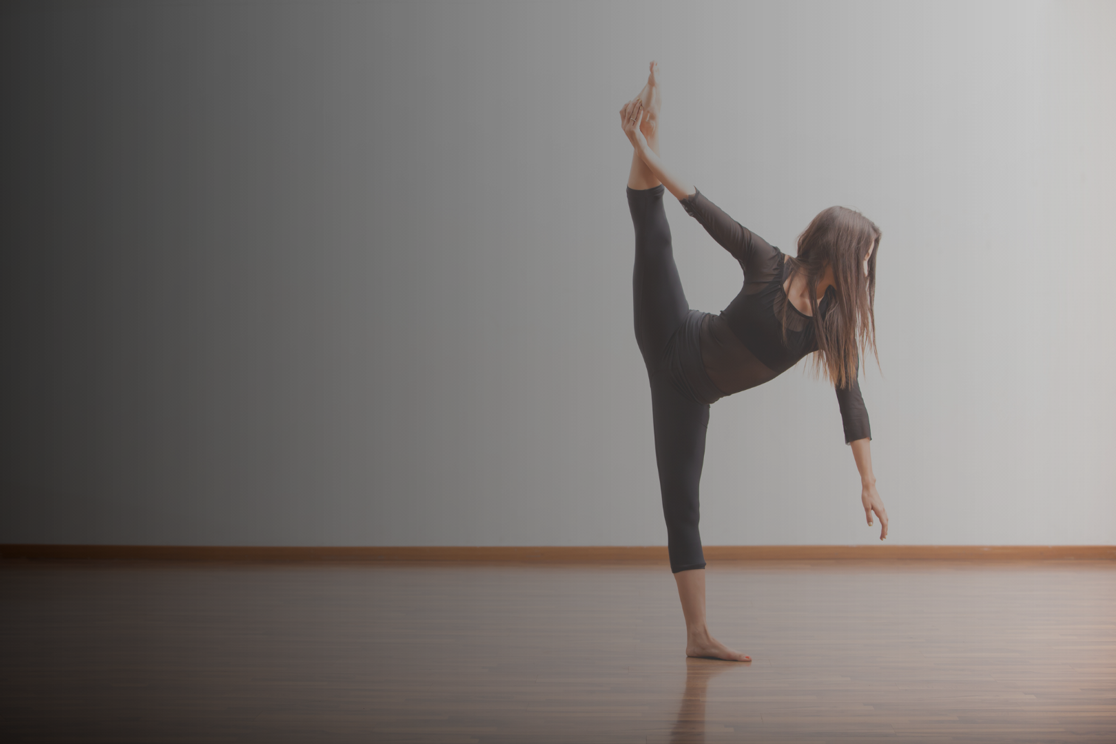 Woman practicing yoga indoors, standing on one leg, with the other leg lifted and held behind her, stretching her arm forward and down.