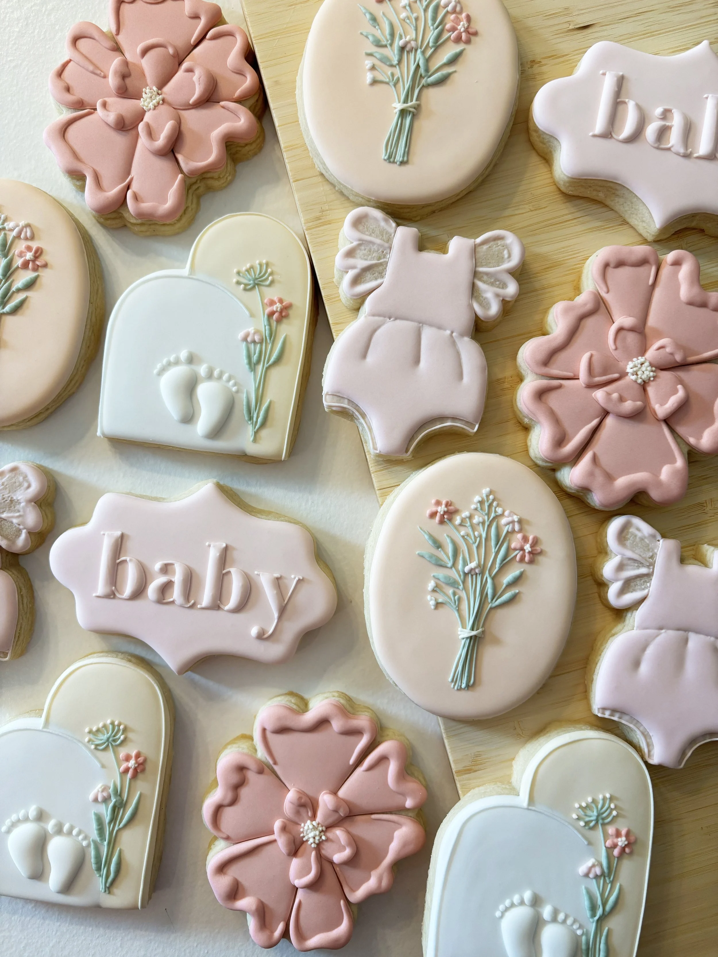 Decorative cookies with pastel icing, featuring baby-themed designs such as bibs, flowers, and the word 'baby' on some cookies.