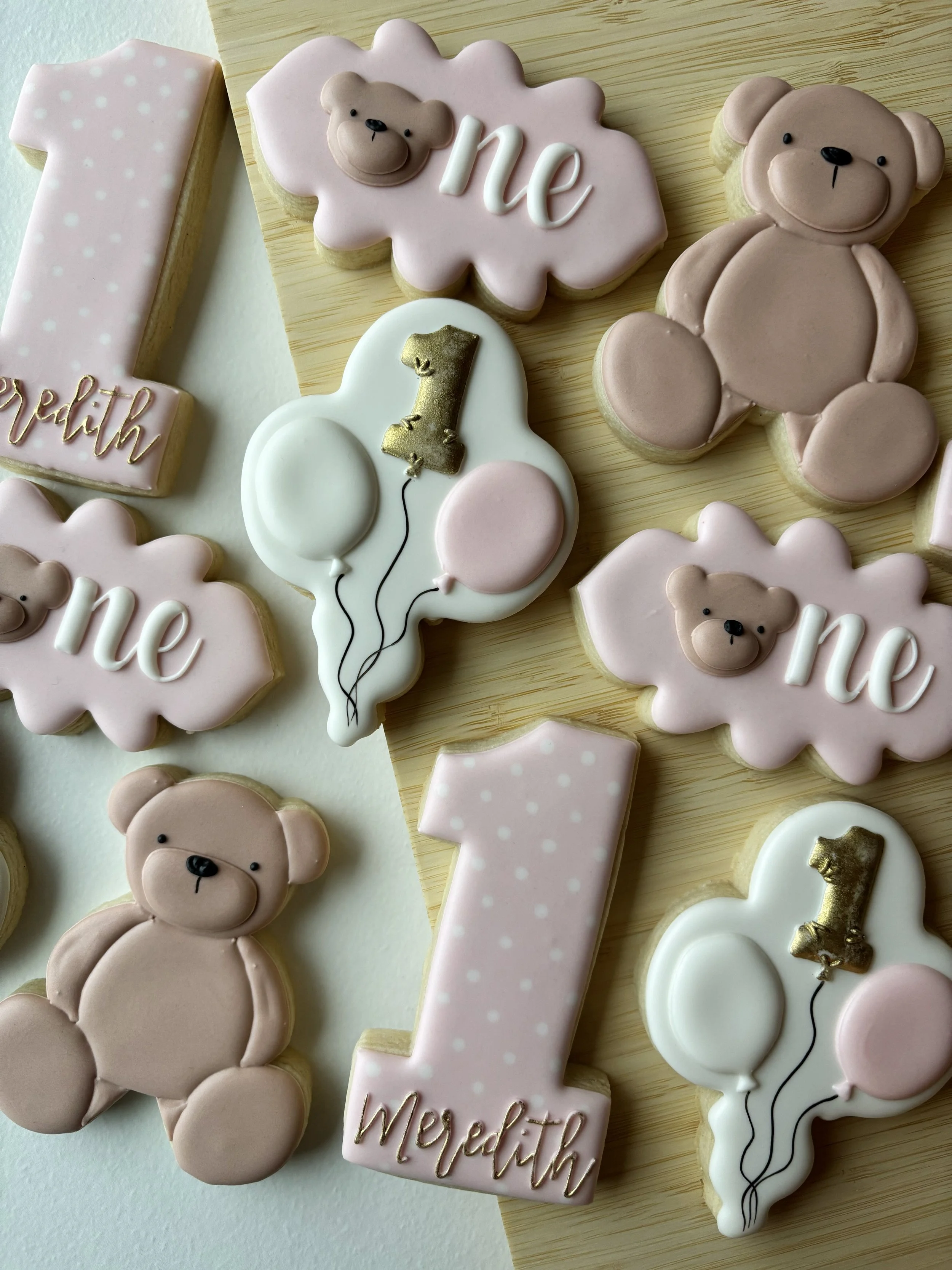 Decorative cookies celebrating a child's first birthday. The cookies include pink teddy bears, pink cloud-shaped cookies with the word "one" and a bear face, large number "1" cookies with "Megan" written below, and balloon-shaped cookies with the num
