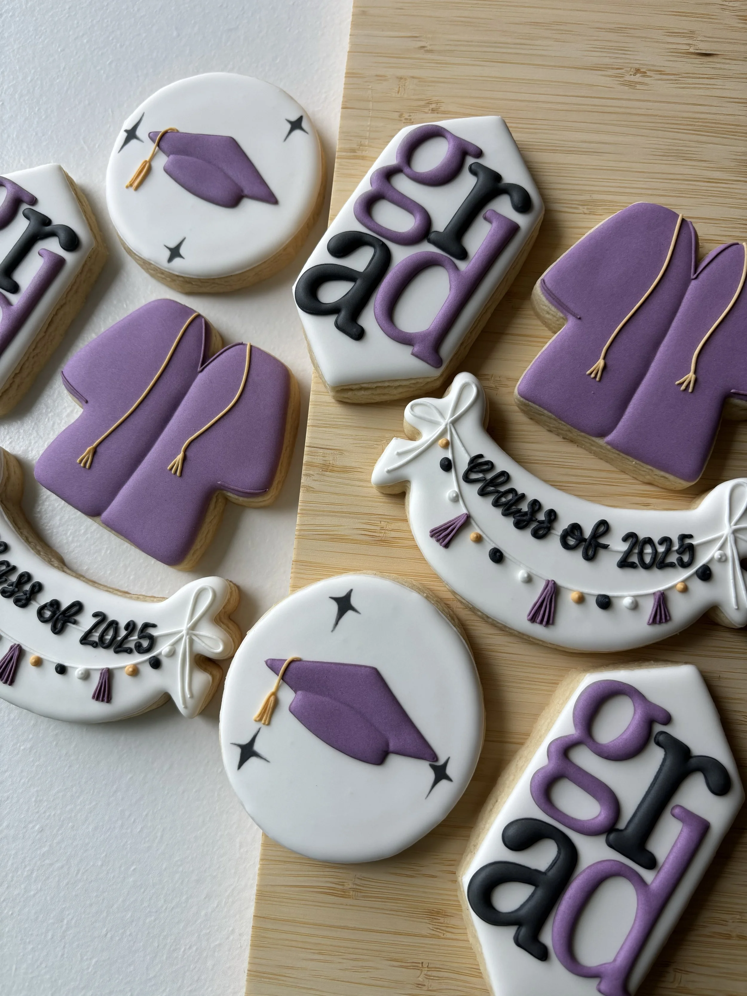 Graduation-themed decorated cookies with purple graduation caps, white scrolls with 'class of 2025' written on them, and purple gown cookies, arranged on a wooden surface.