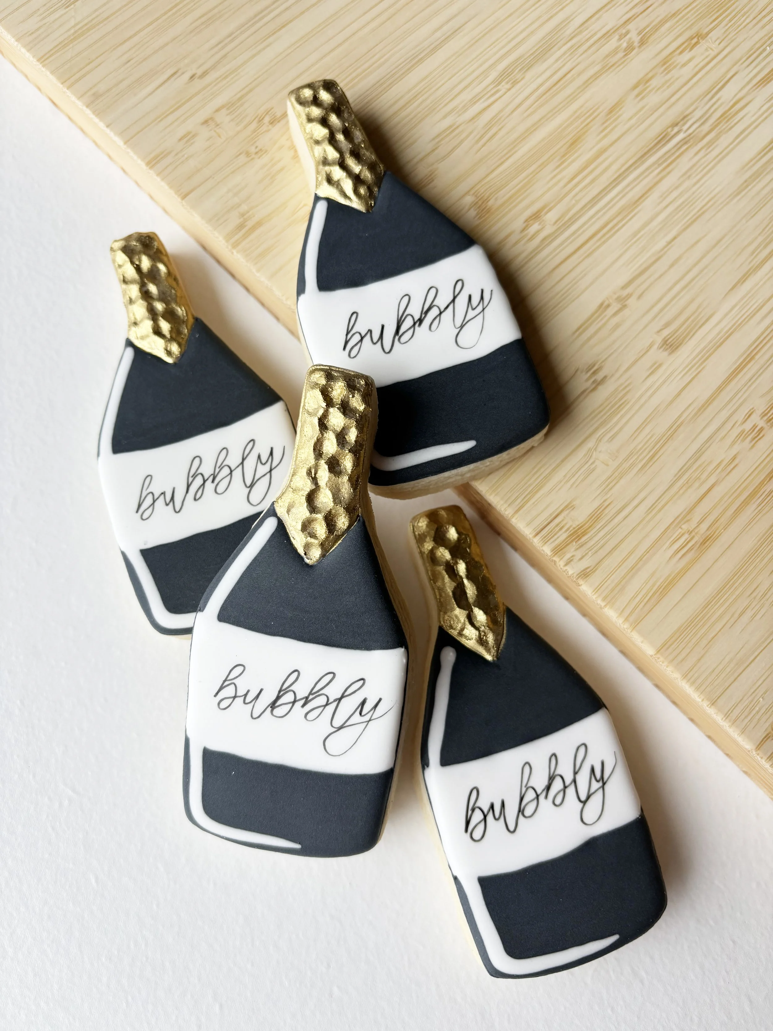 Decorative cookies shaped like champagne bottles with black, white, and gold details, with the word 'bubbly' written on them.