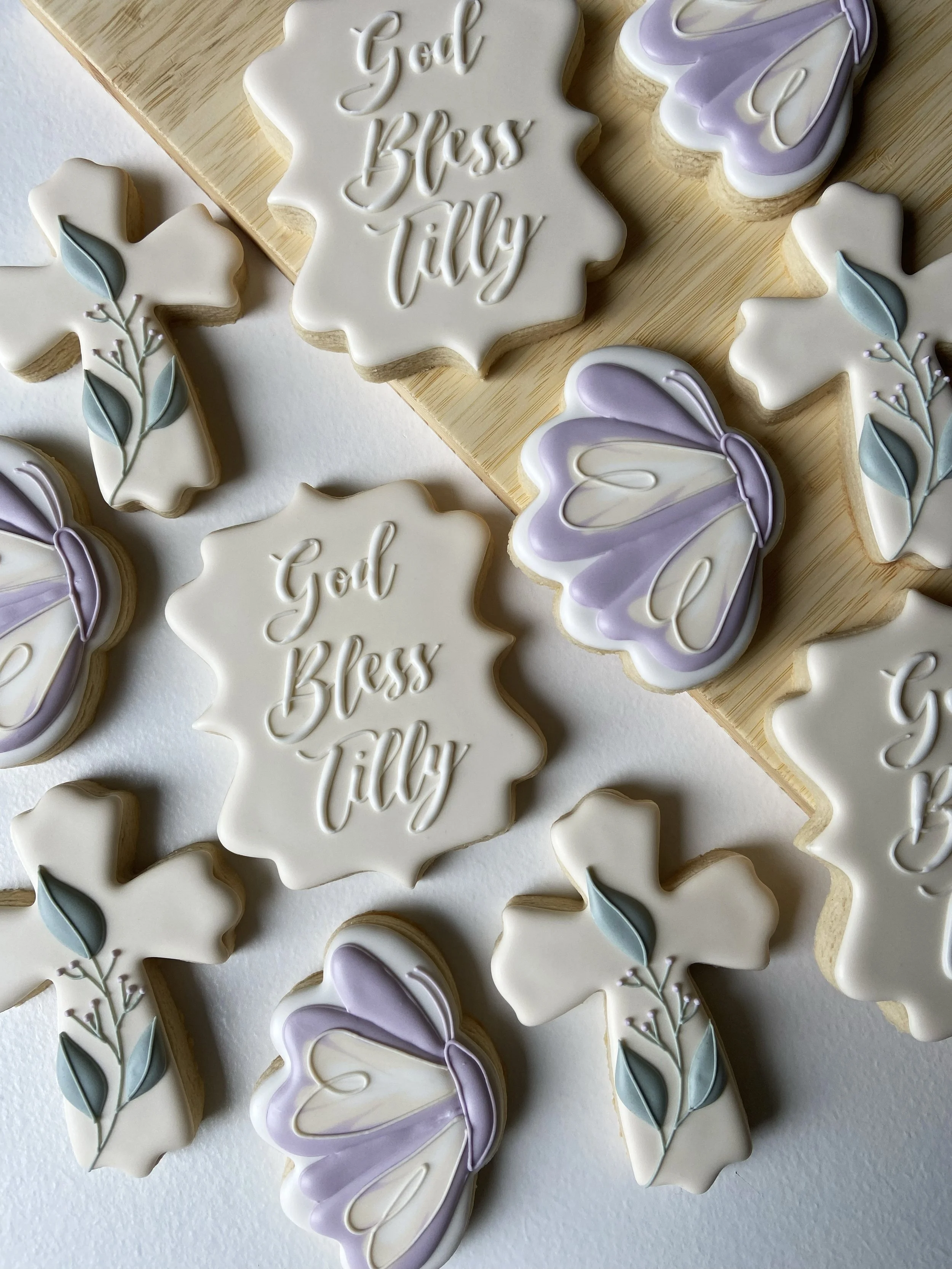 Decorated sugar cookies with religious and floral designs, some with the words "God Bless Tilly" and others with purple butterfly and floral decorations.