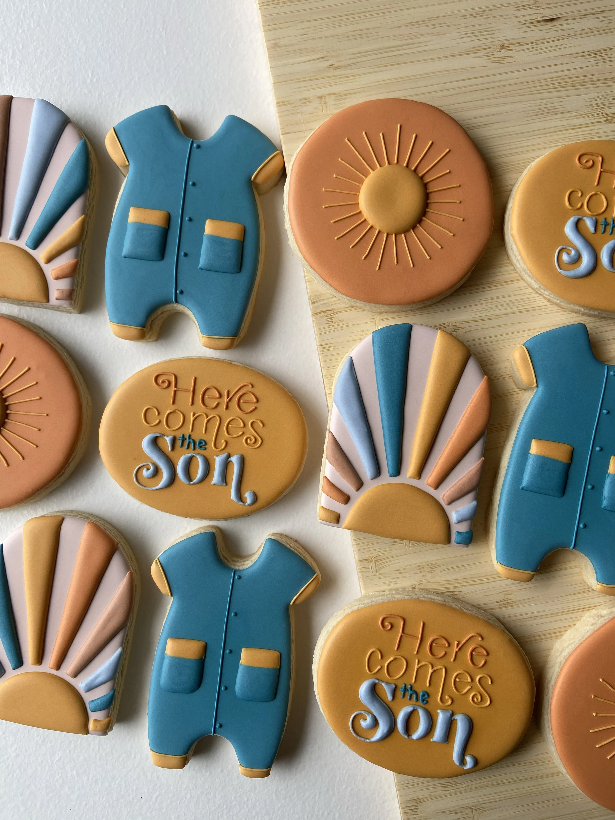 Decorative cookies themed for a celebration, featuring sun designs, sunrise illustrations, baby clothes, and the text "Here comes the son".
