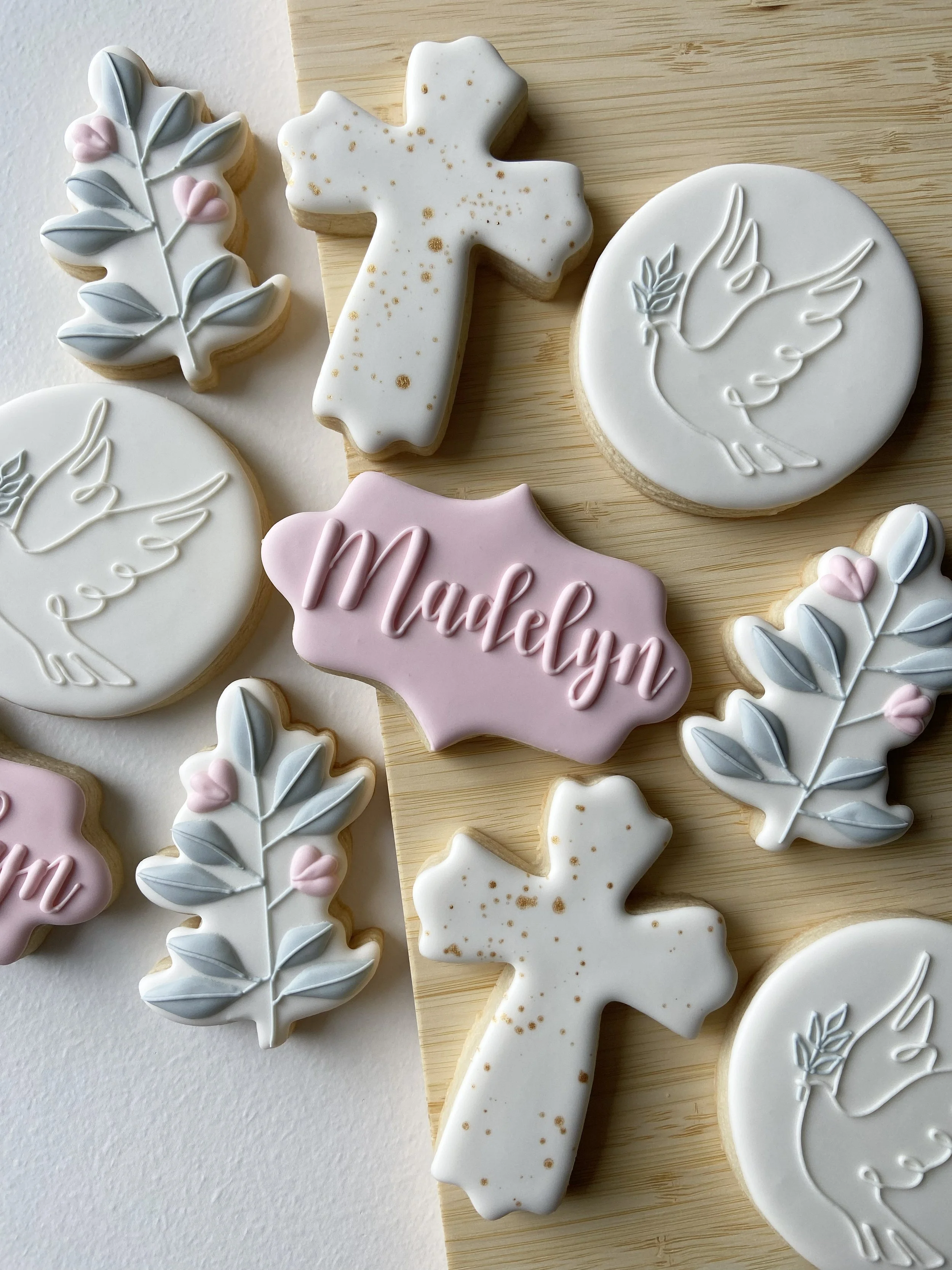 Decorative cookies shaped like crosses, leaves, and circles, with pastel icing in white, pink, and blue, some featuring embossed bird and leaf designs and one with the name 'Madalyn' written in pink lettering.