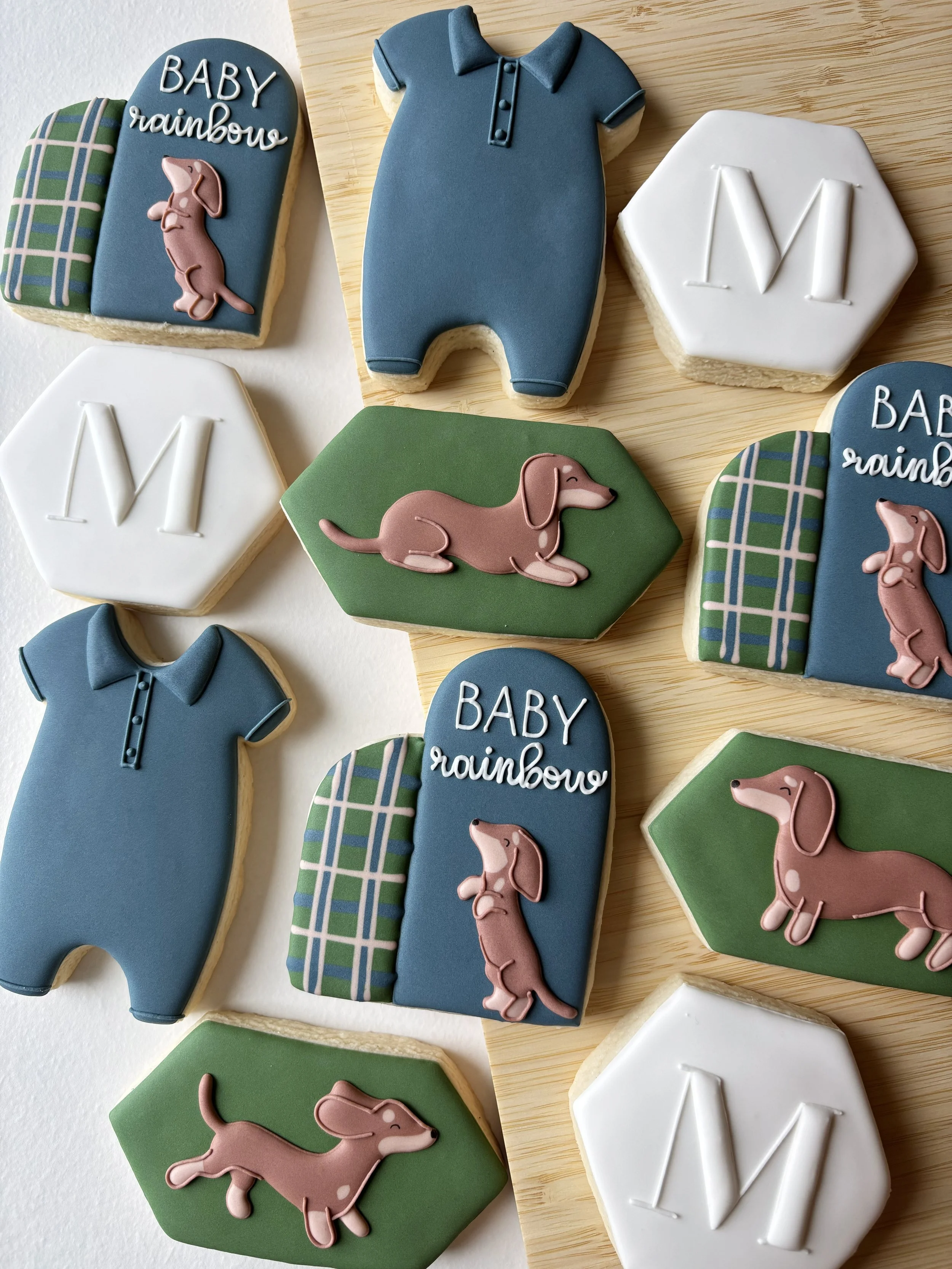 Collection of decorated cookies with baby shower theme, including ones shaped like dachshund dogs, green hexagons, white hexagons with letter M, and blue ones with baby rainbow and plaid patterns.