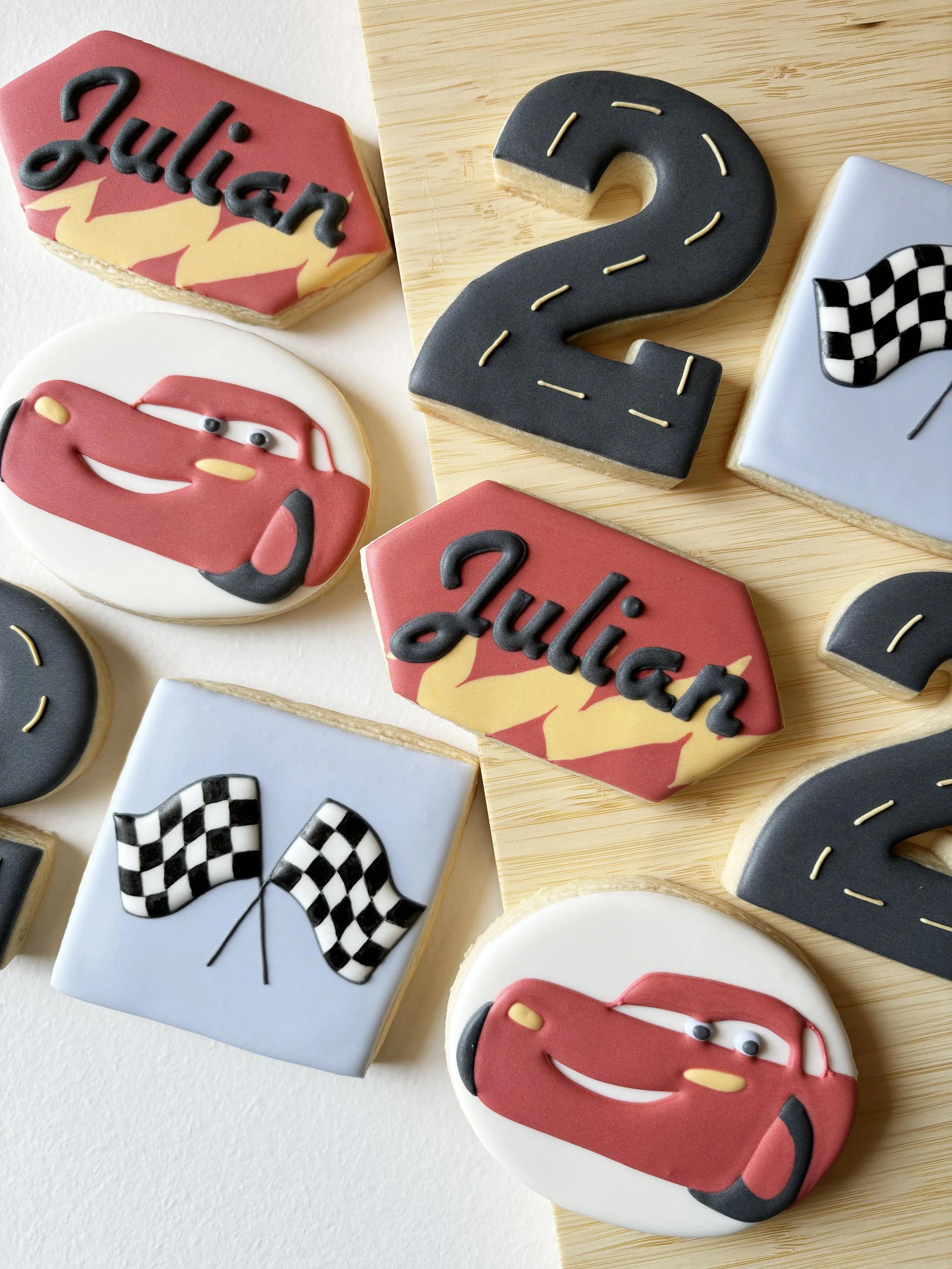Assorted decorated cookies themed to racing, featuring checkered flags, a car face, the number "2" with yellow lines, and the name "Julian" in black script on red and yellow backgrounds, arranged on a light wooden surface.