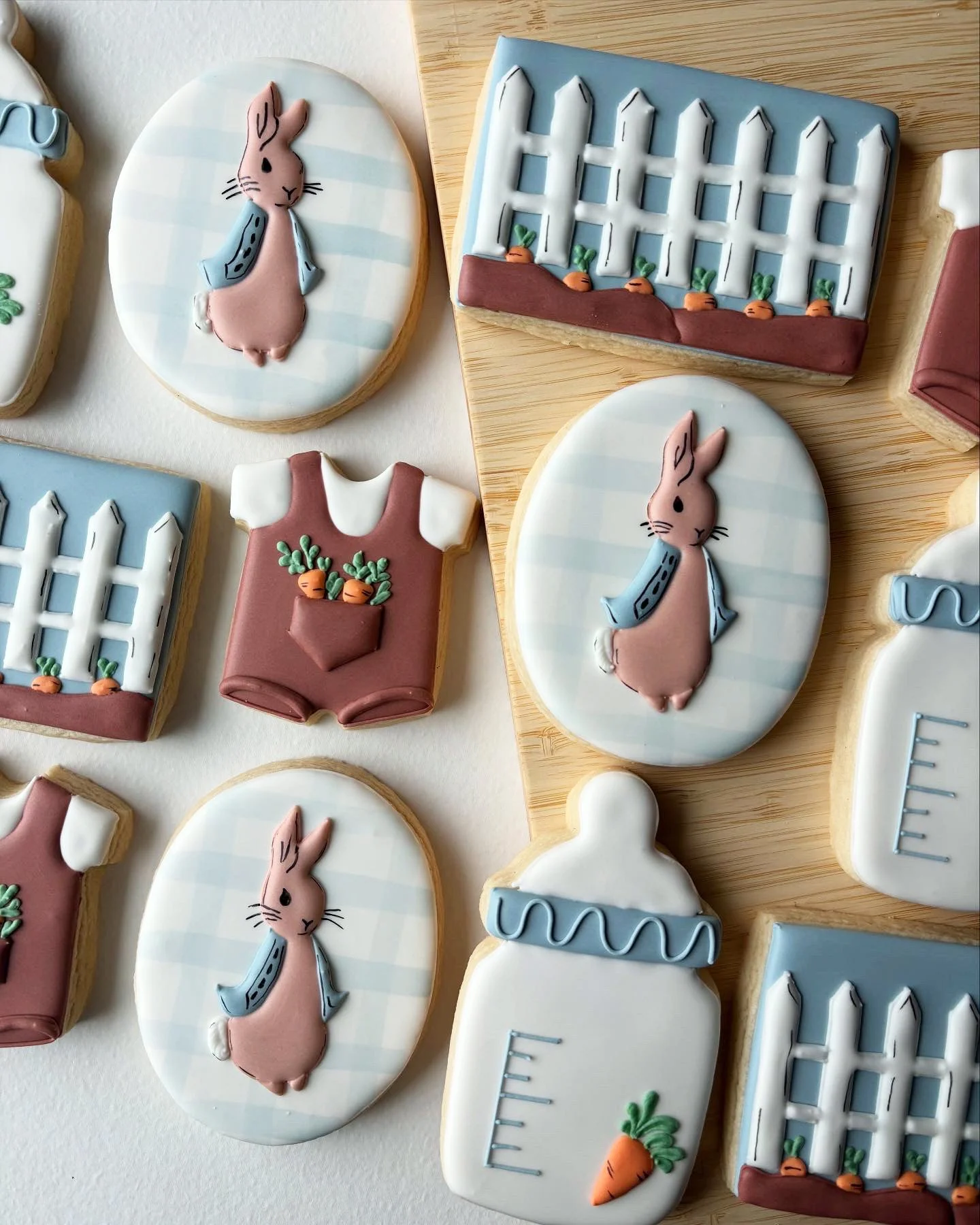 Assorted Easter-themed decorated cookies featuring bunnies, carrots, baby onesies with carrots, and picket fences, on a wooden surface.
