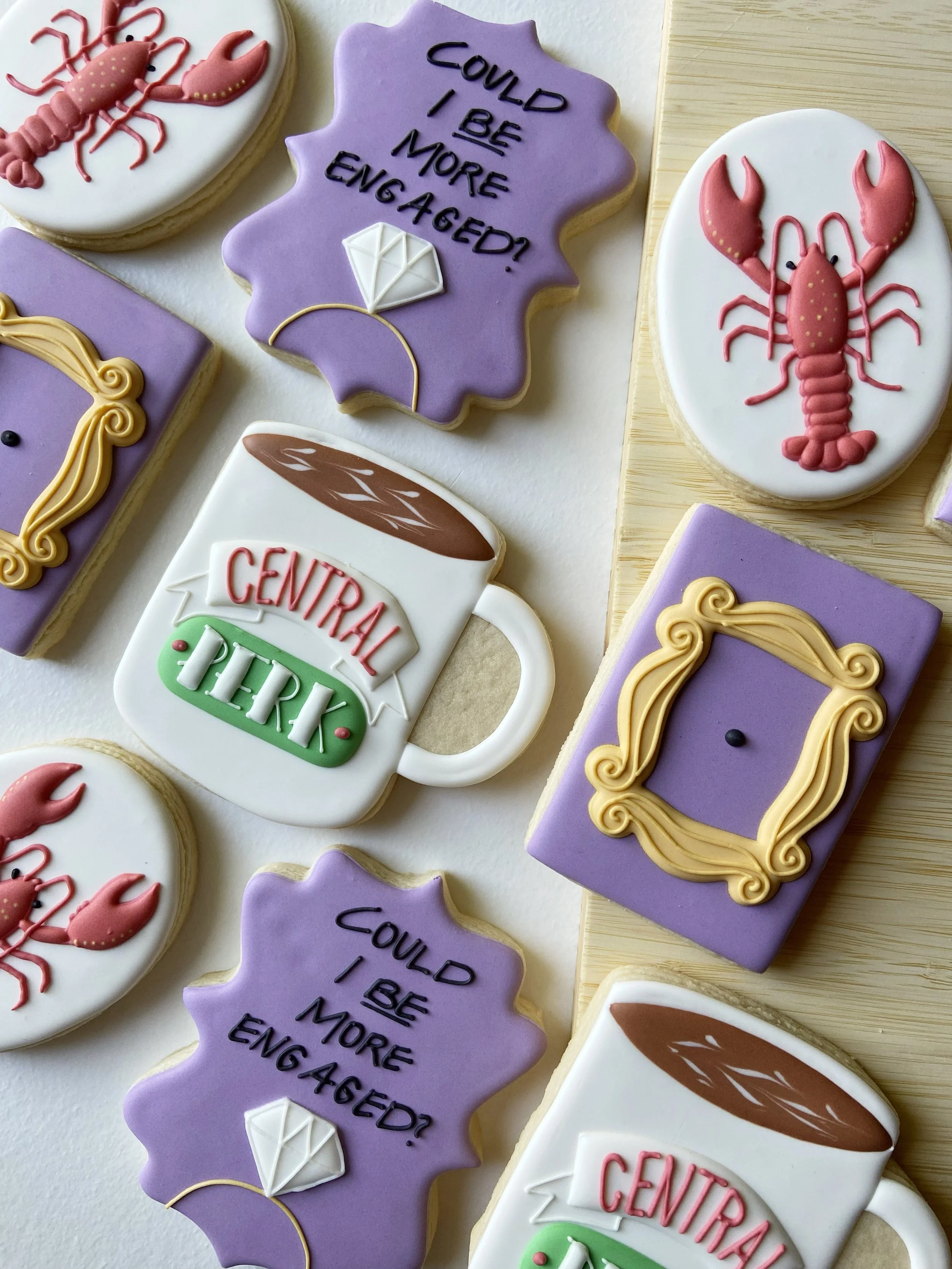 Decorative cookies with various designs including some with a lobster, a diamond, a coffee mug with 'Central Perk' written on it, and an empty picture frame, all in pastel colors.