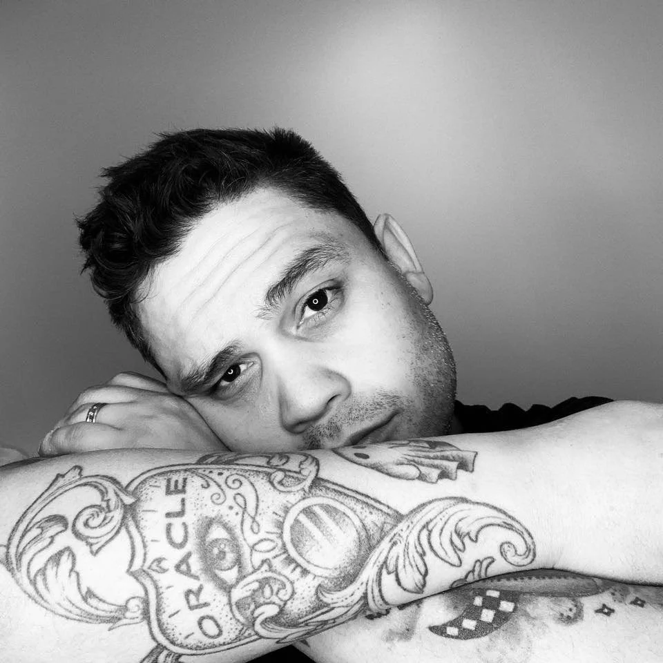 Black and white photo of a man resting his head on his arm, looking into the camera. He has short dark hair, facial stubble, and a tattoo on his forearm that reads 'ORACLE' with decorative designs.