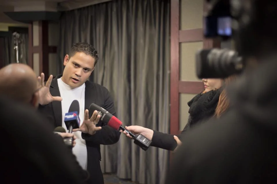 A man being interviewed by multiple reporters, with microphones, in an indoor setting with curtains in the background.