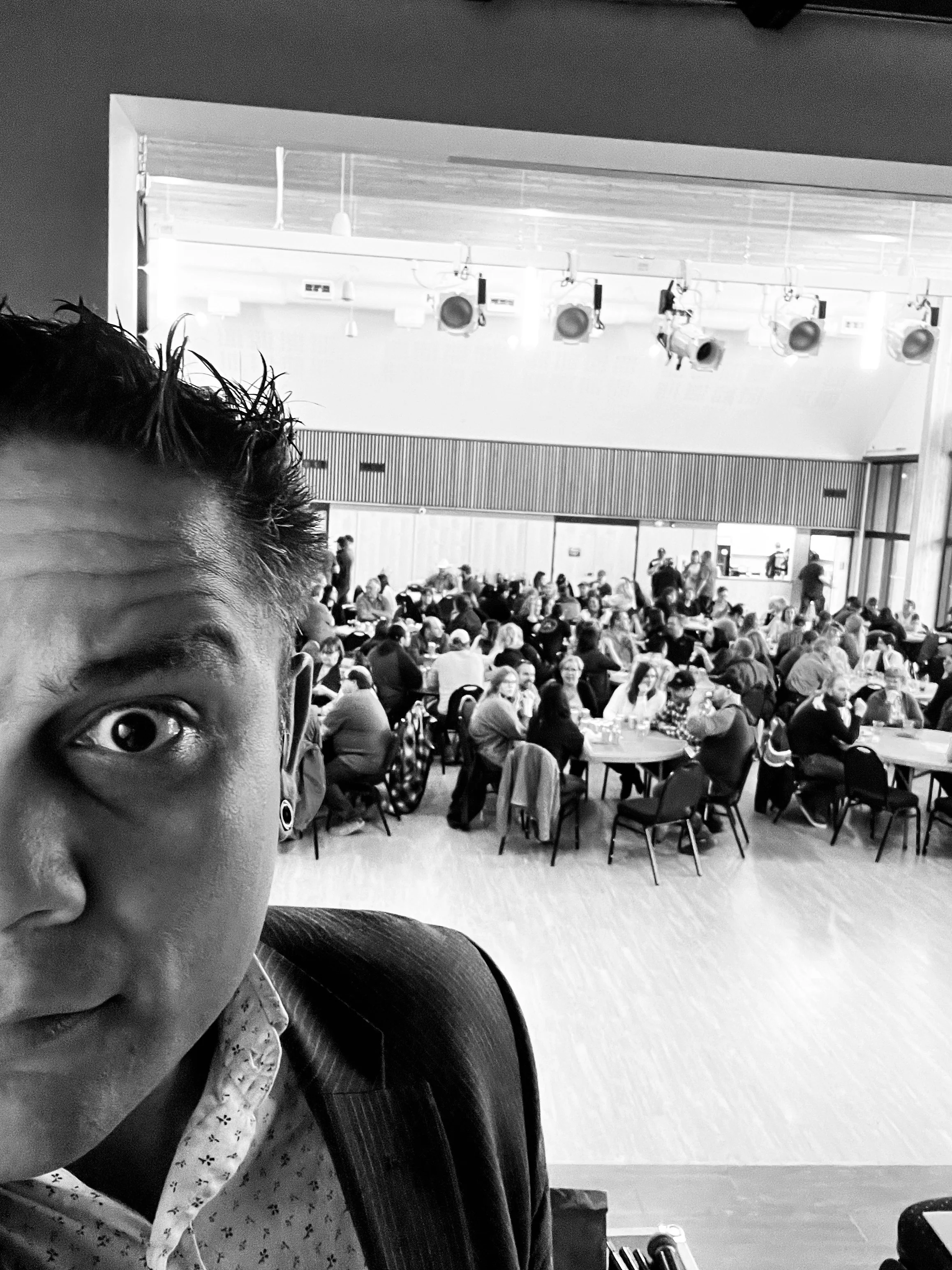 Black and white photo of man in foreground with a busy event hall background filled with people sitting at tables.