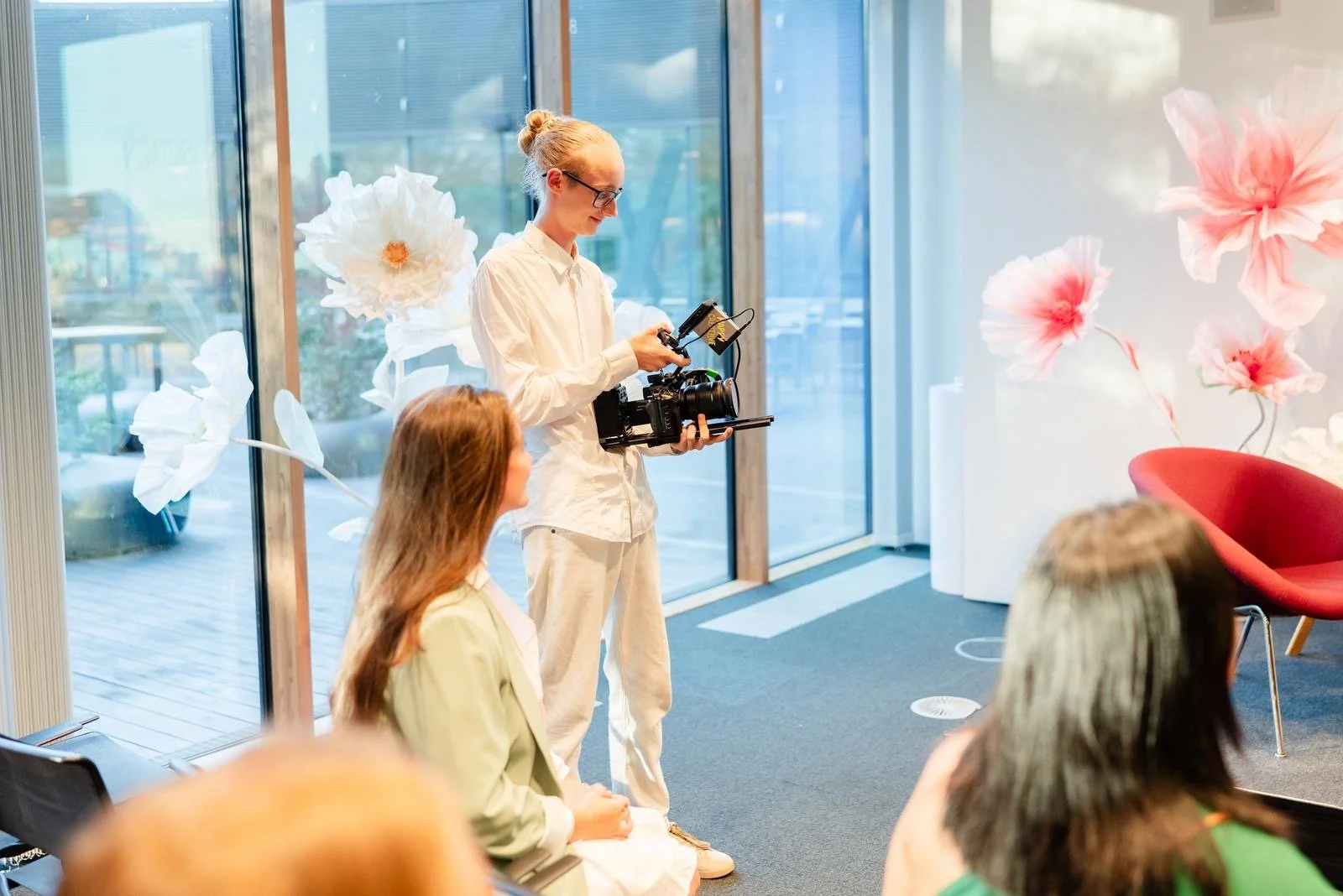 Eine Frau mit Brille hält eine Kamera vor einem weißen Hintergrund mit großen, weißen und pinken Papierblumen. Vor ihr sitzen drei Frauen, die einer Präsentation oder einem Gespräch folgen.