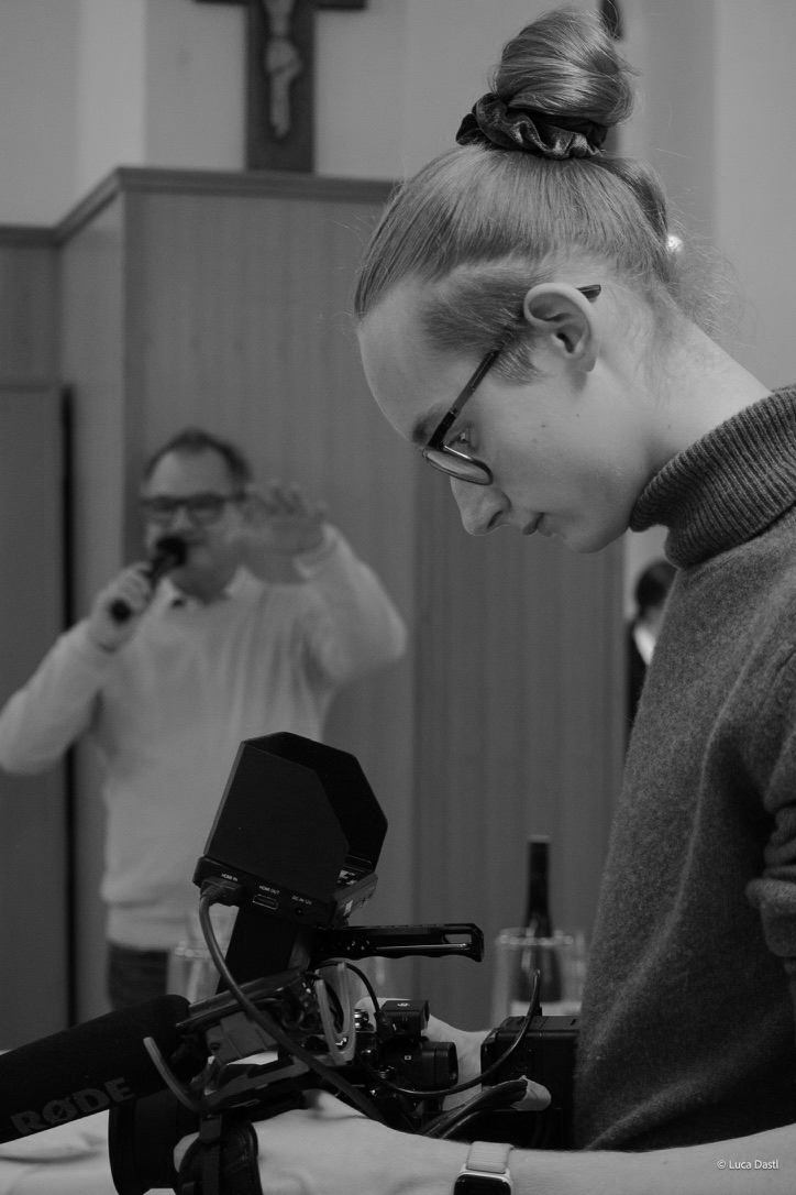 Eine junge Frau mit Brille und hochgestecktem Haar arbeitet an einer Kamera, im Hintergrund ein Mann mit Bart, der eine Flasche hält und an einem Mikrofon nippt.