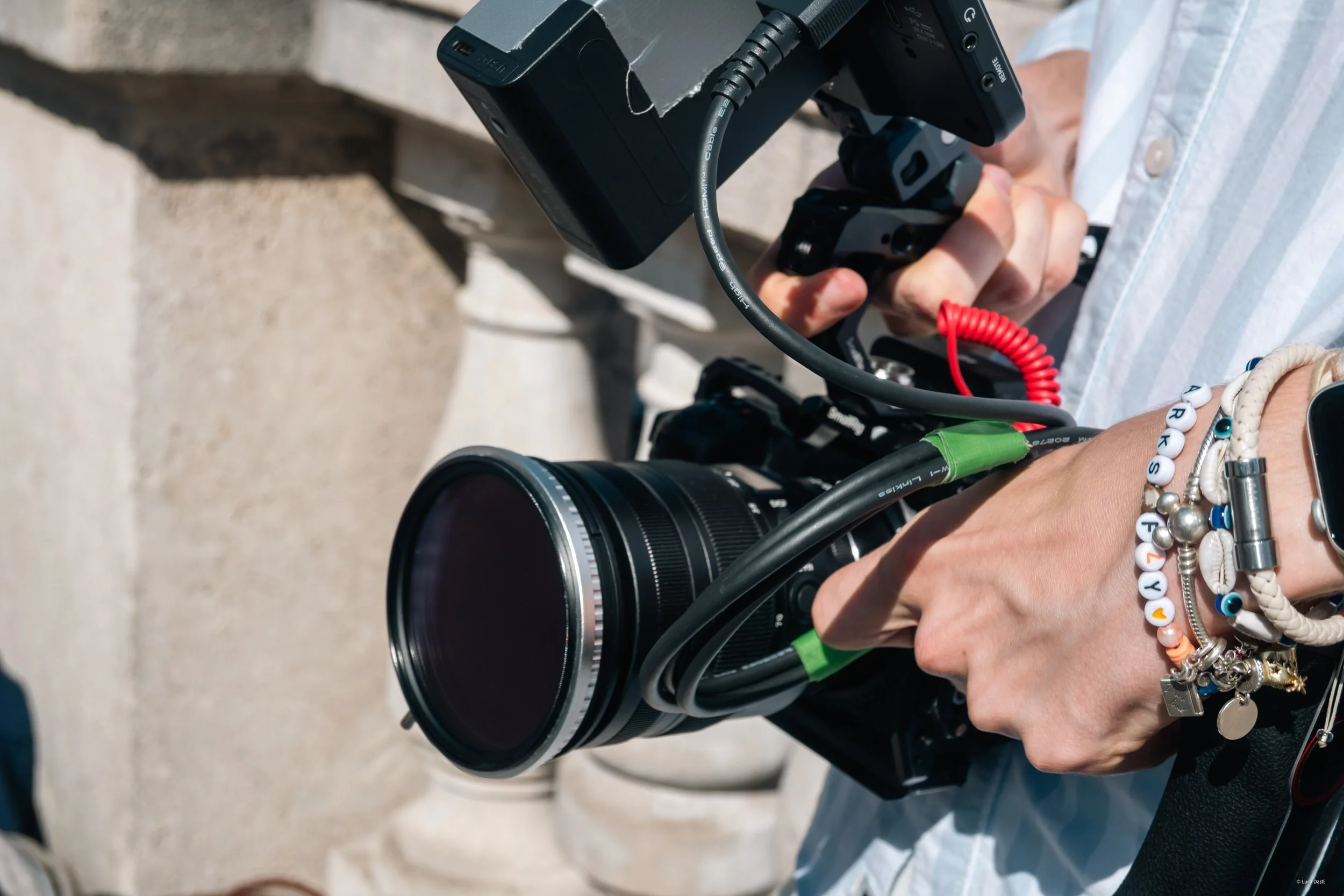Nahaufnahme einer Kamera, die von einer Person gehalten wird, mit mehreren Armbändern am Handgelenk, vor einem hellen Steinhintergrund.
