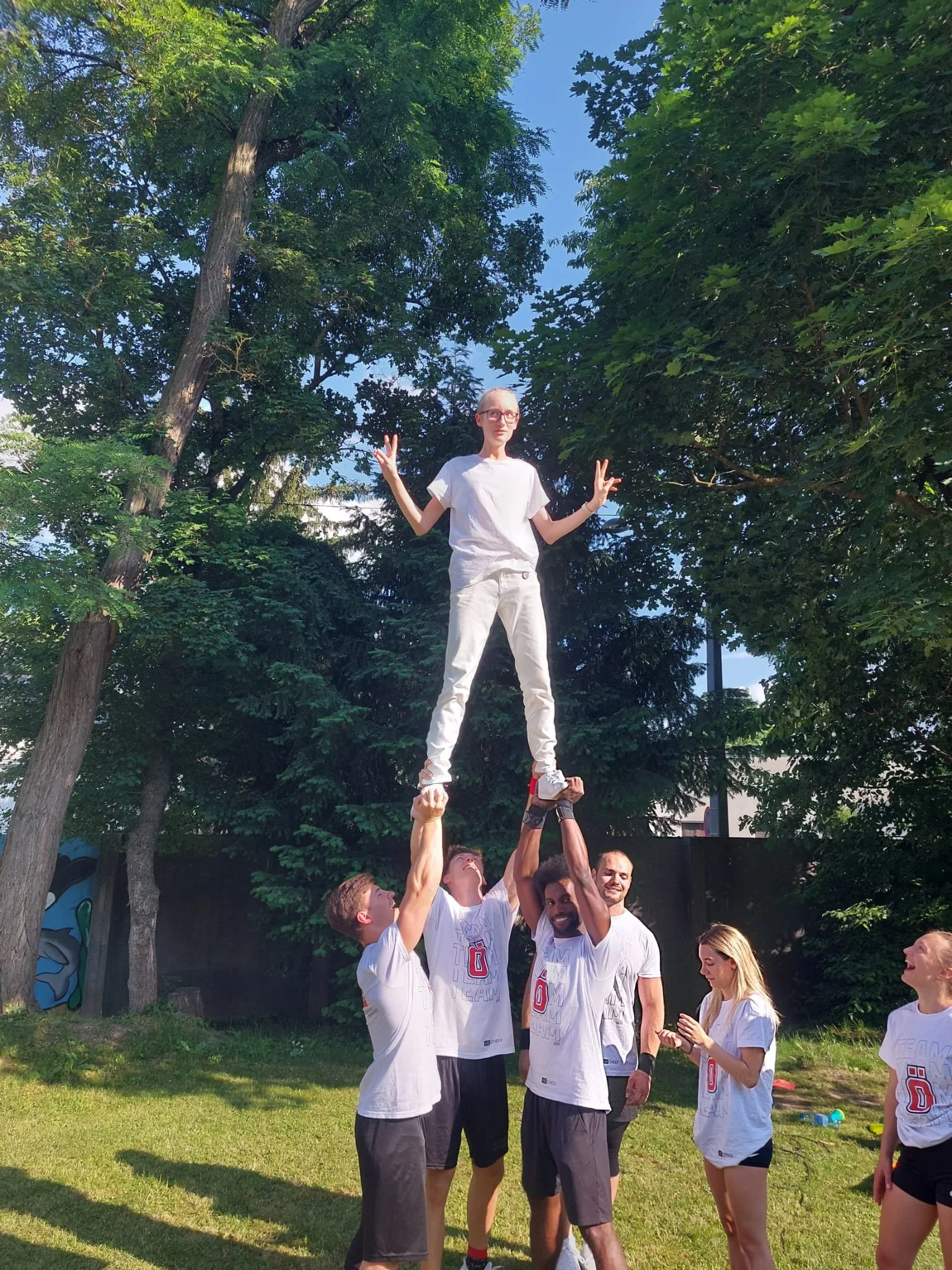 Eine Gruppe junger Menschen macht mit einem Balanceakt im Freien Spaß. Einige stützen einen Jungen, der auf den Händen zweier anderer steht, während andere zuschauen und Fotos machen. Das Ganze findet bei Tageslicht vor Bäumen statt.